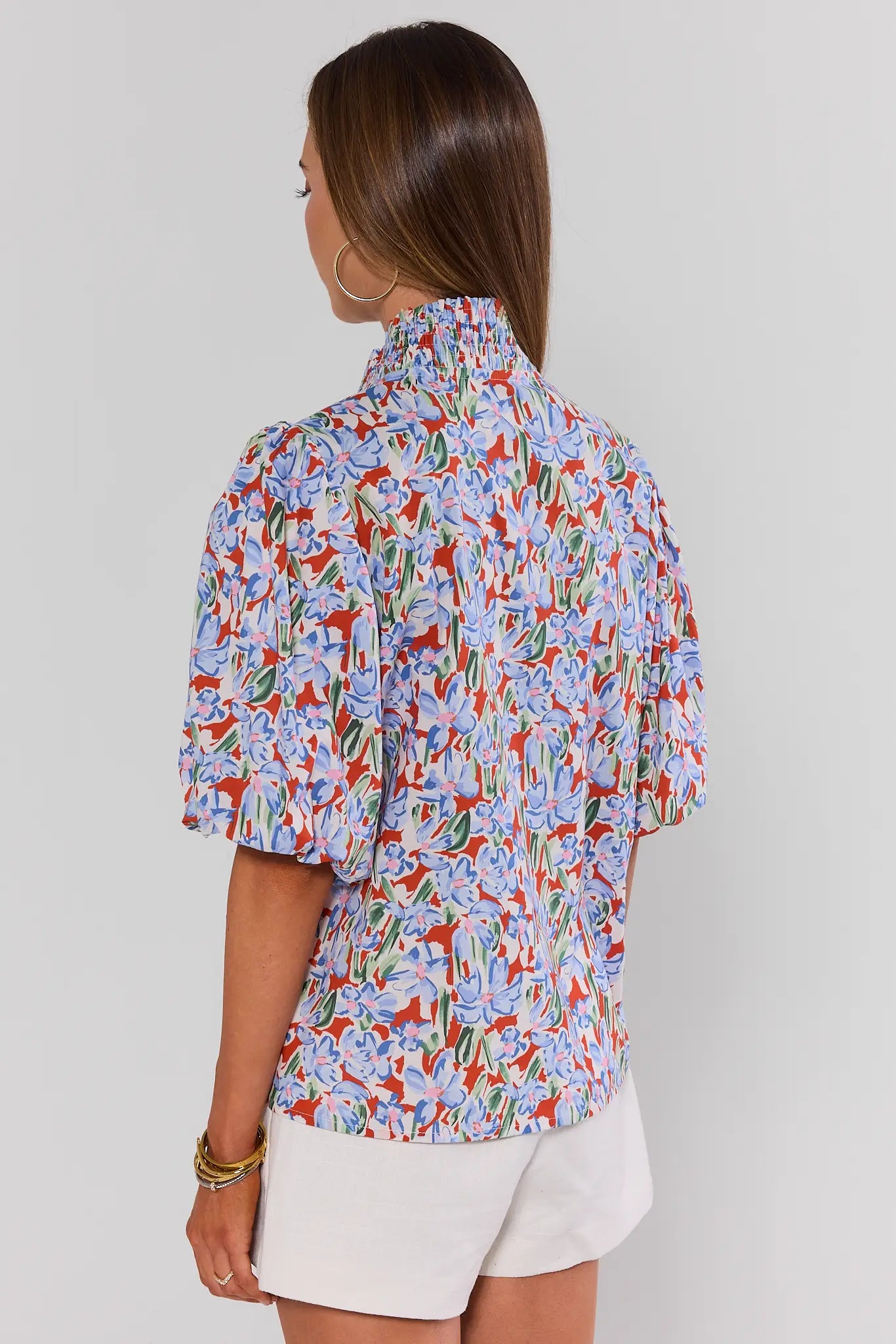 Woman standing wearing a colorful floral blouse and white shorts on a plain background