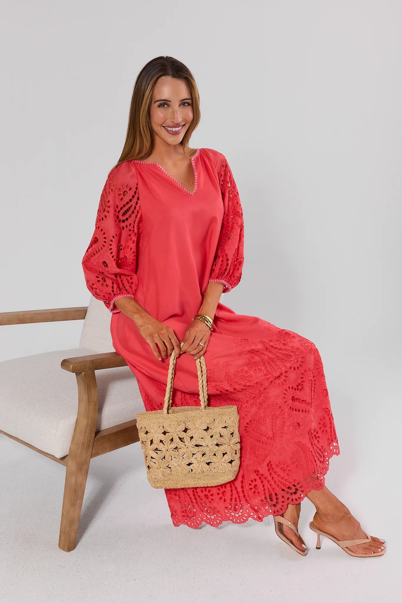 Woman in a coral dress holding a woven bag, sitting on a white chair with a white background