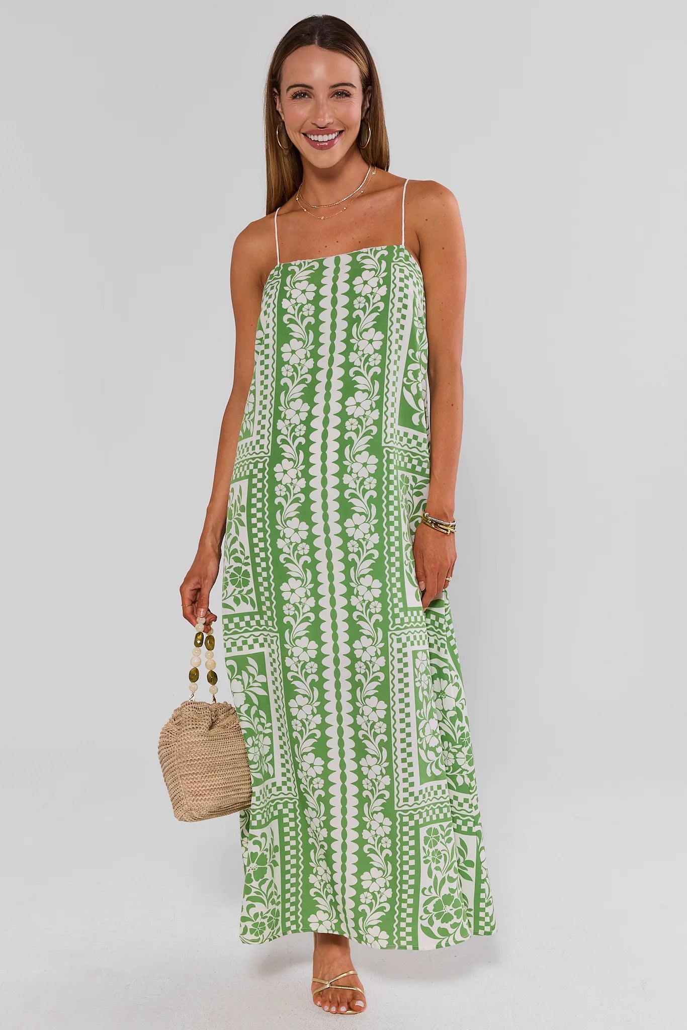 Woman wearing a green and white patterned dress holding a straw bag on a light gray background