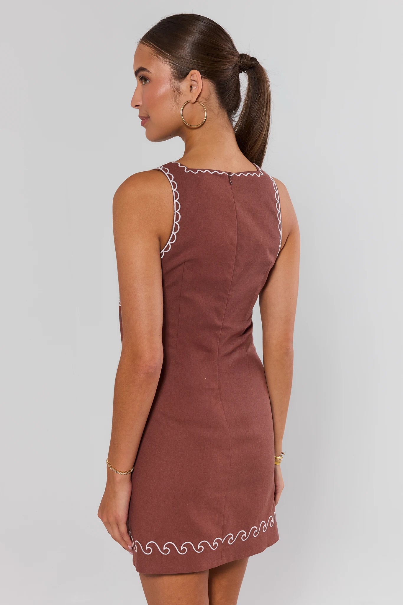 Woman standing wearing a brown dress with white trim on a plain background