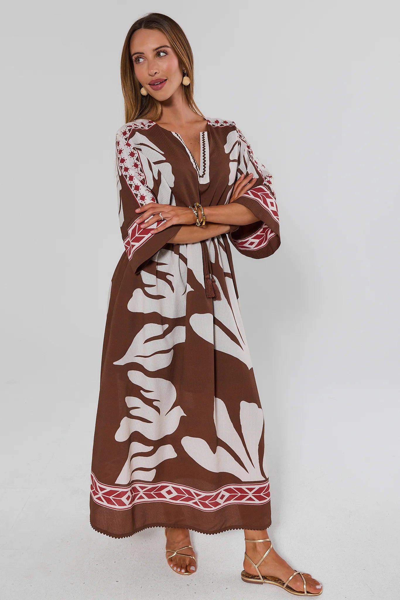Woman wearing a brown and white patterned dress with red accents on a plain background with arms crossed