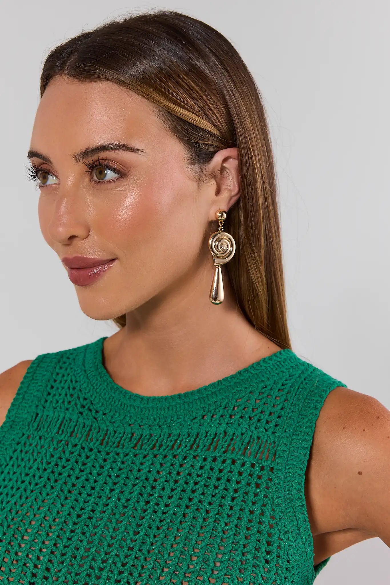 Woman wearing a green sleeveless top and gold shell earrings with a plain background