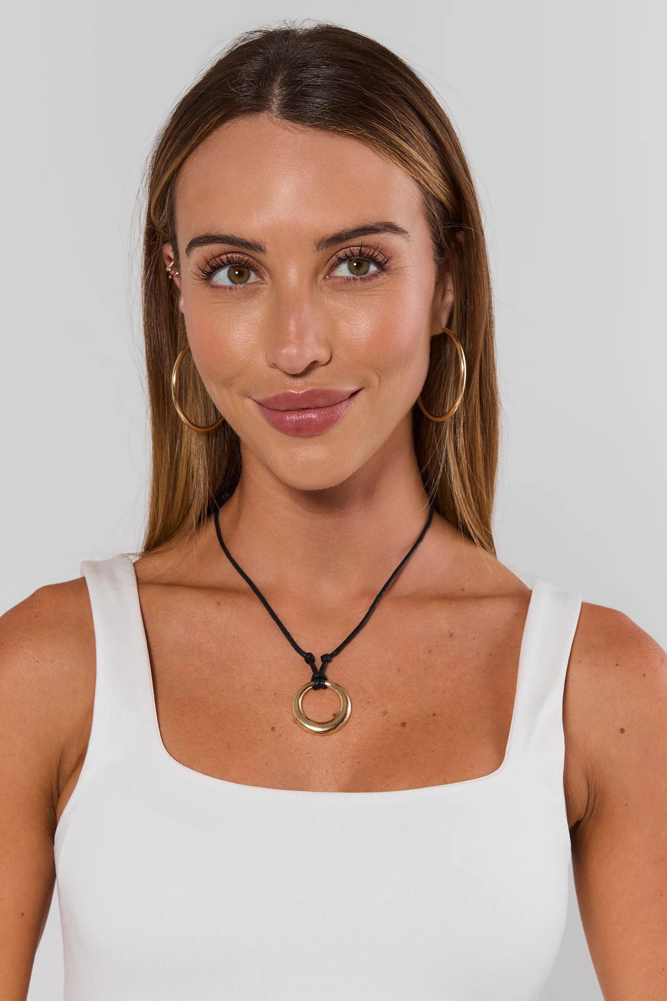 Woman wearing a white tank top and a black necklace with a gold pendant against a plain background