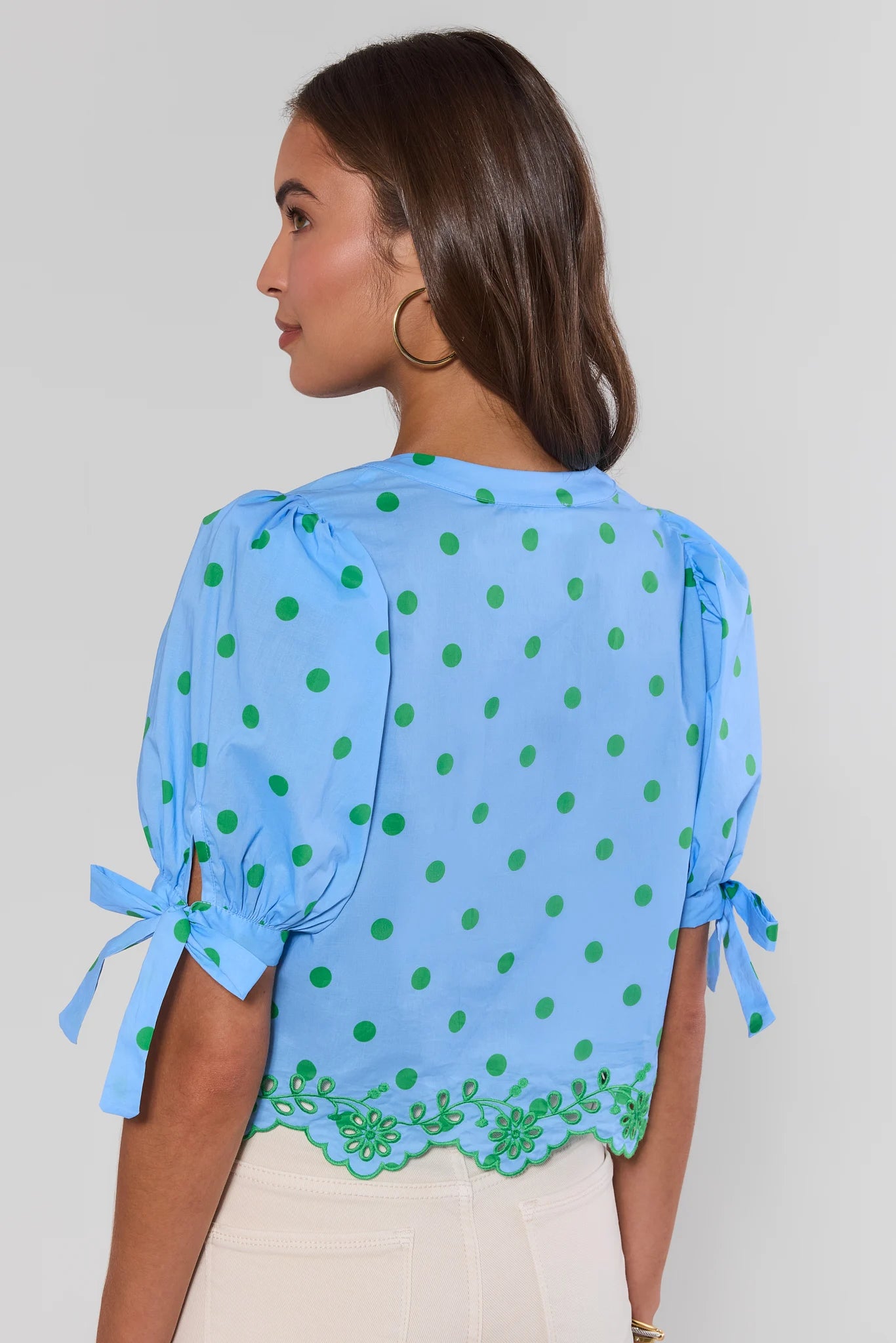 Woman standing wearing a blue blouse with green polka dots and lace trim on a plain background