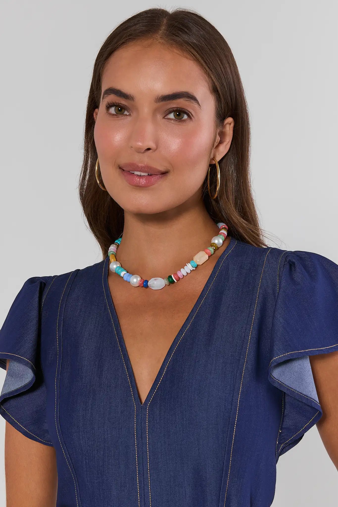 Woman wearing a blue dress with ruffled sleeves and a colorful beaded necklace on a plain background