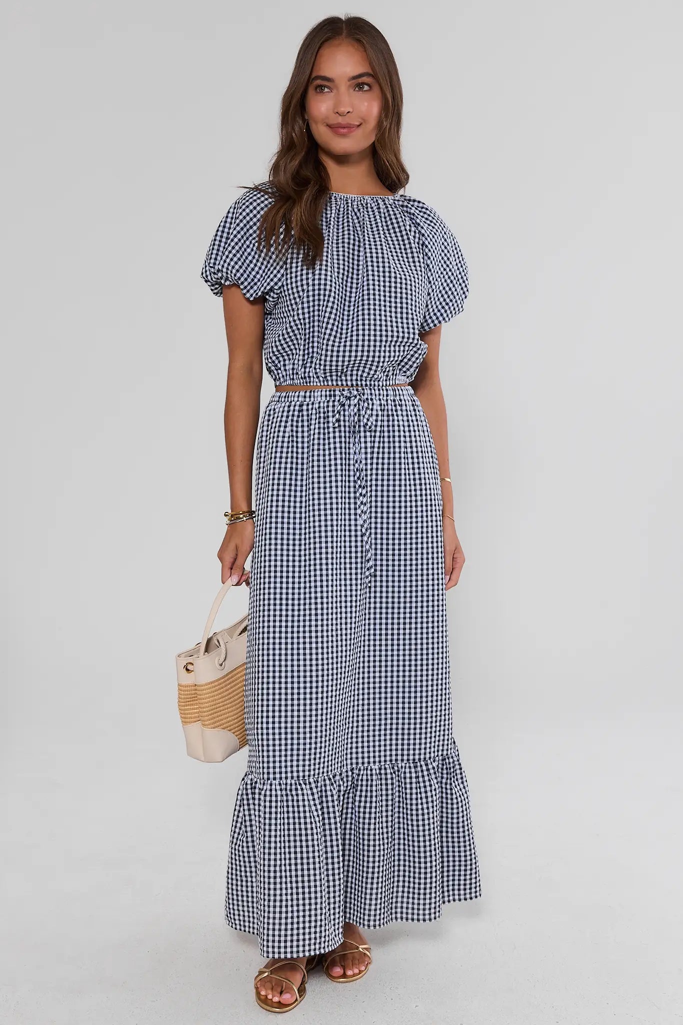 Woman wearing a blue and white checkered top and skirt holding a woven bag on a white background
