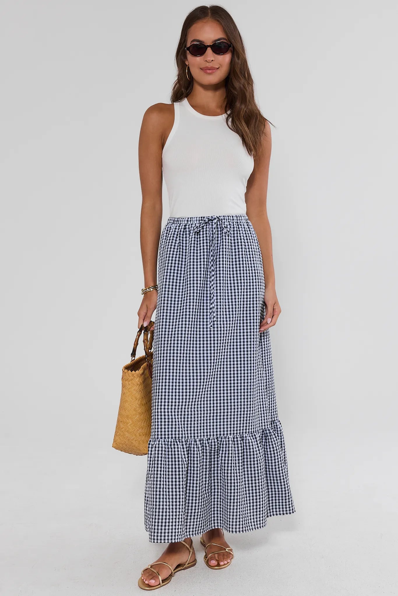 Woman wearing a white sleeveless top and blue checkered skirt, holding a straw bag on a white background