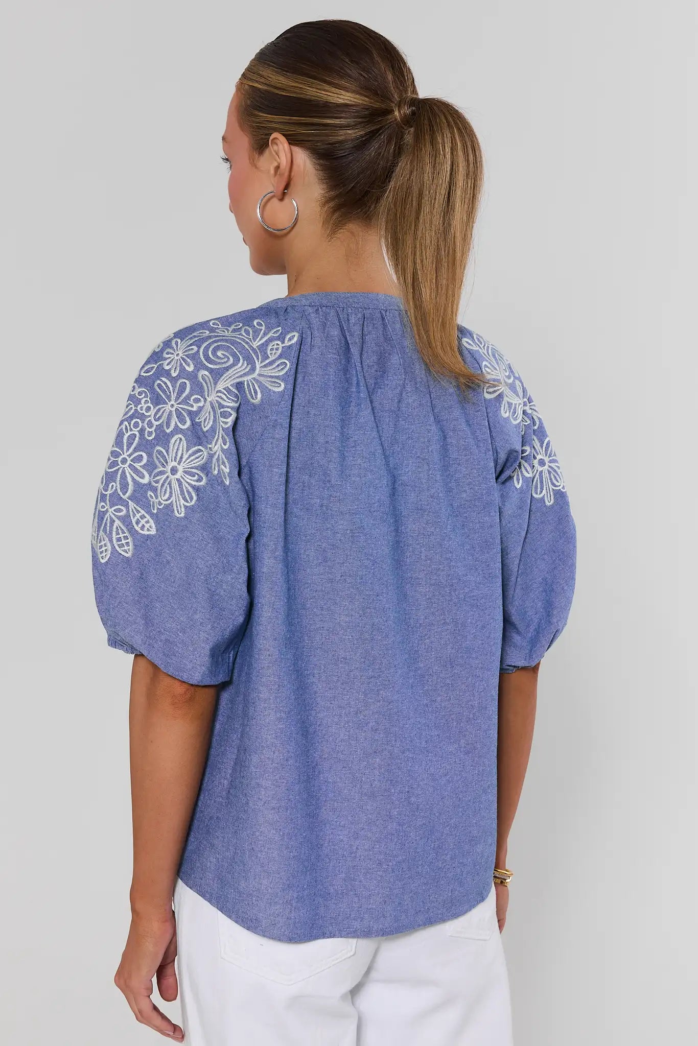 Woman wearing a blue blouse with white floral embroidery on a plain background standing