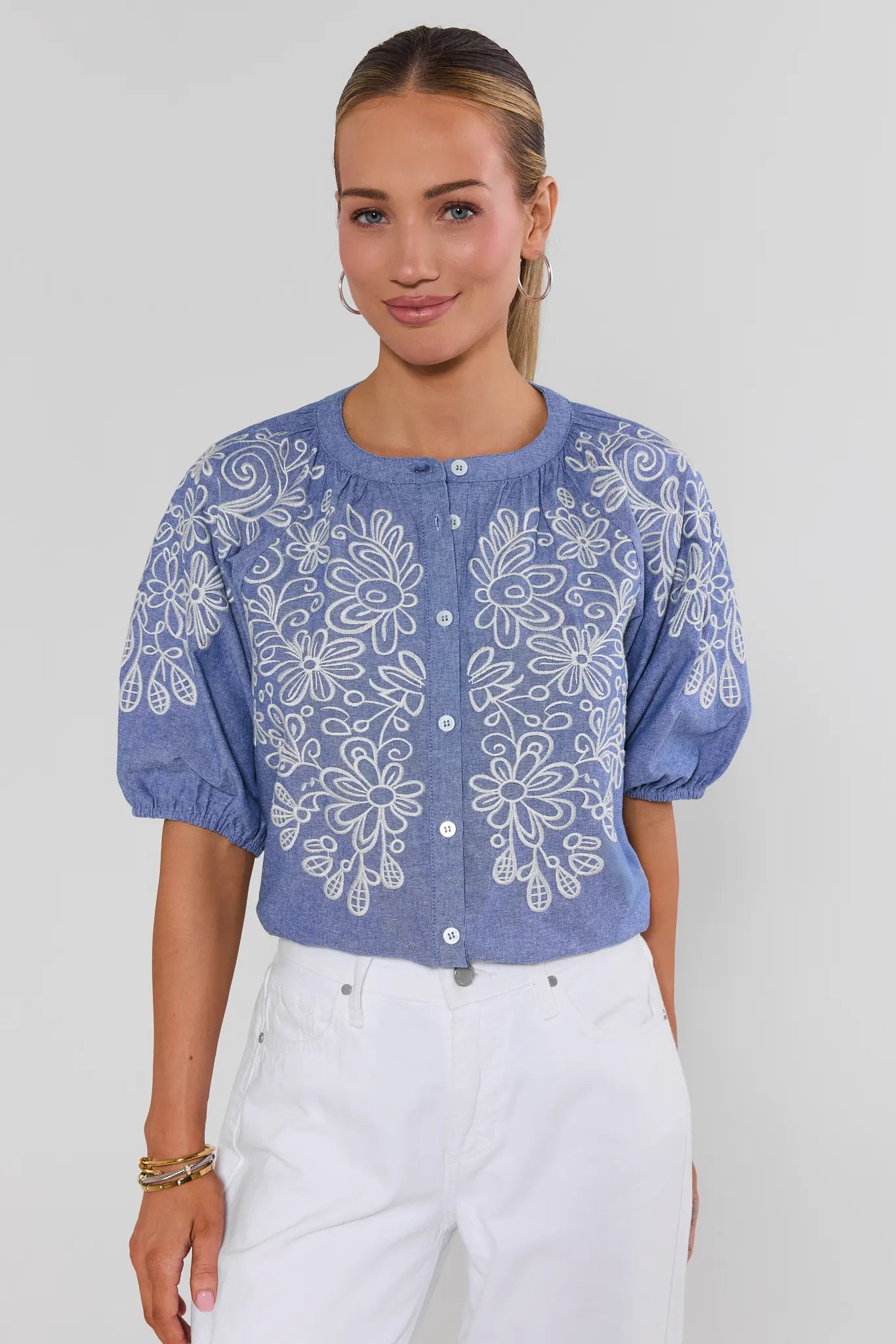 Woman wearing a blue embroidered blouse with white pants on a plain background