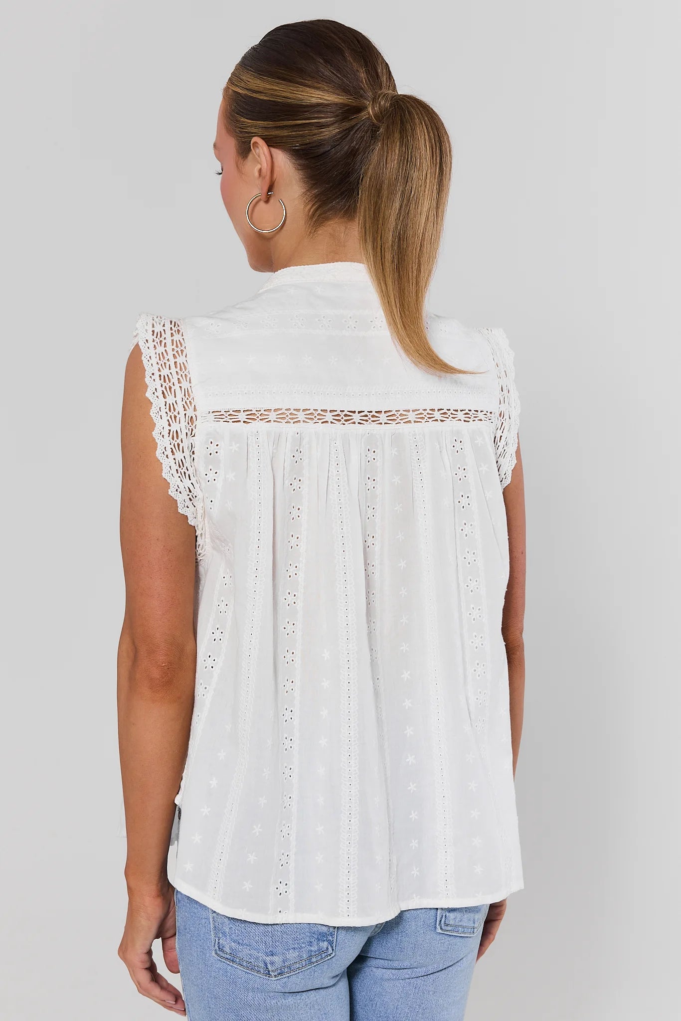 Woman wearing a white sleeveless top with lace details on a plain background stand