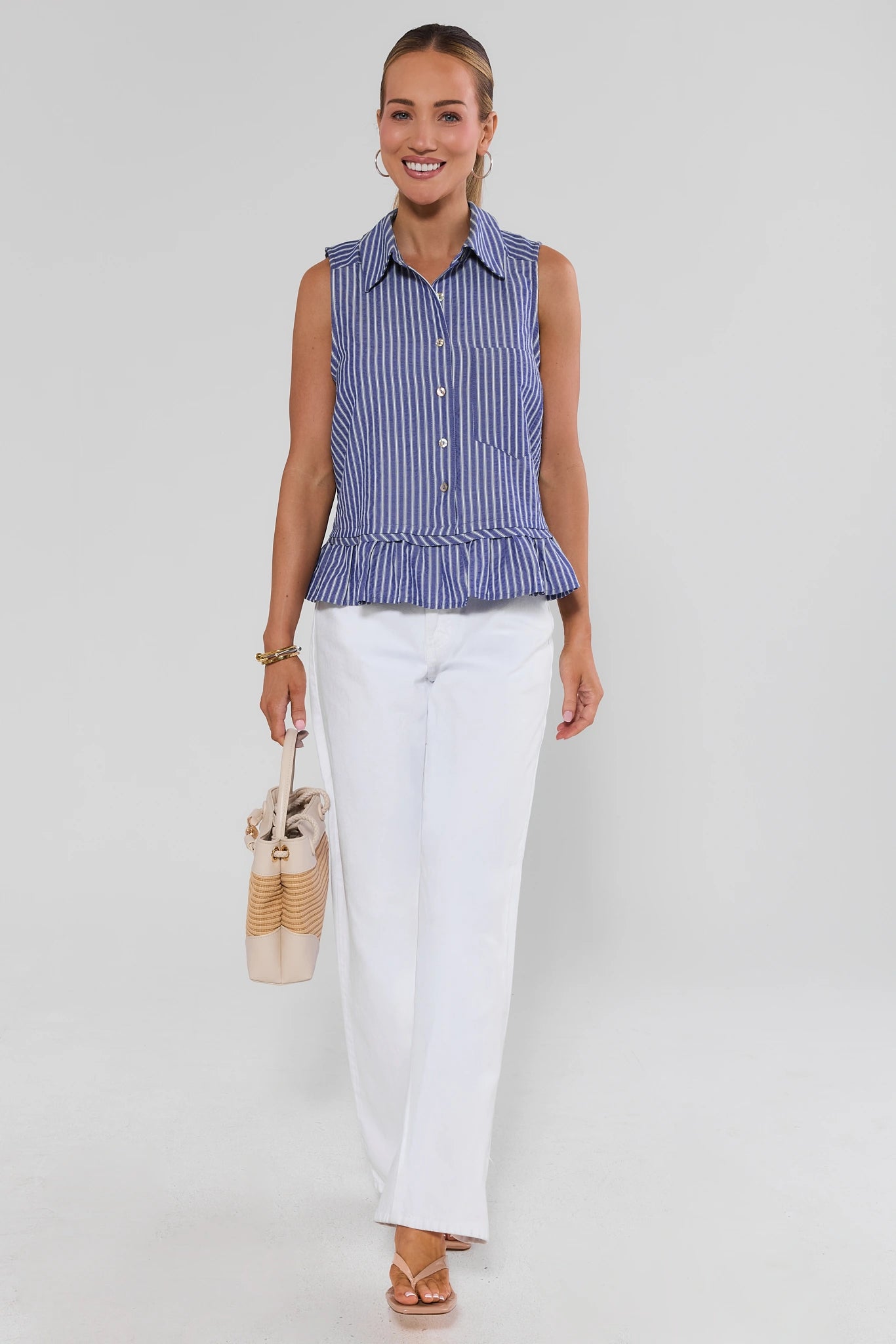 Woman wearing a blue striped sleeveless top and white pants on a light gray background walking