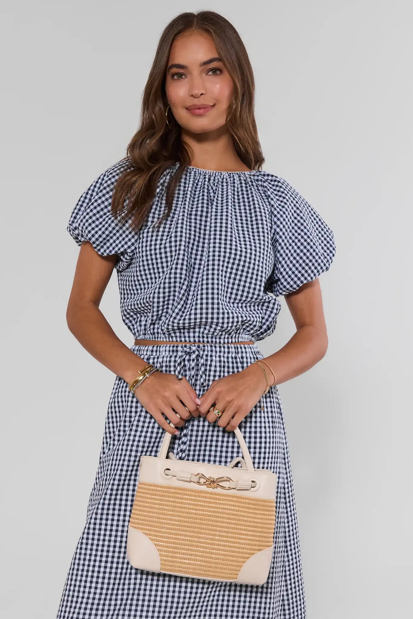 Woman wearing a blue and white checkered dress holding a beige and white handbag on a plain background