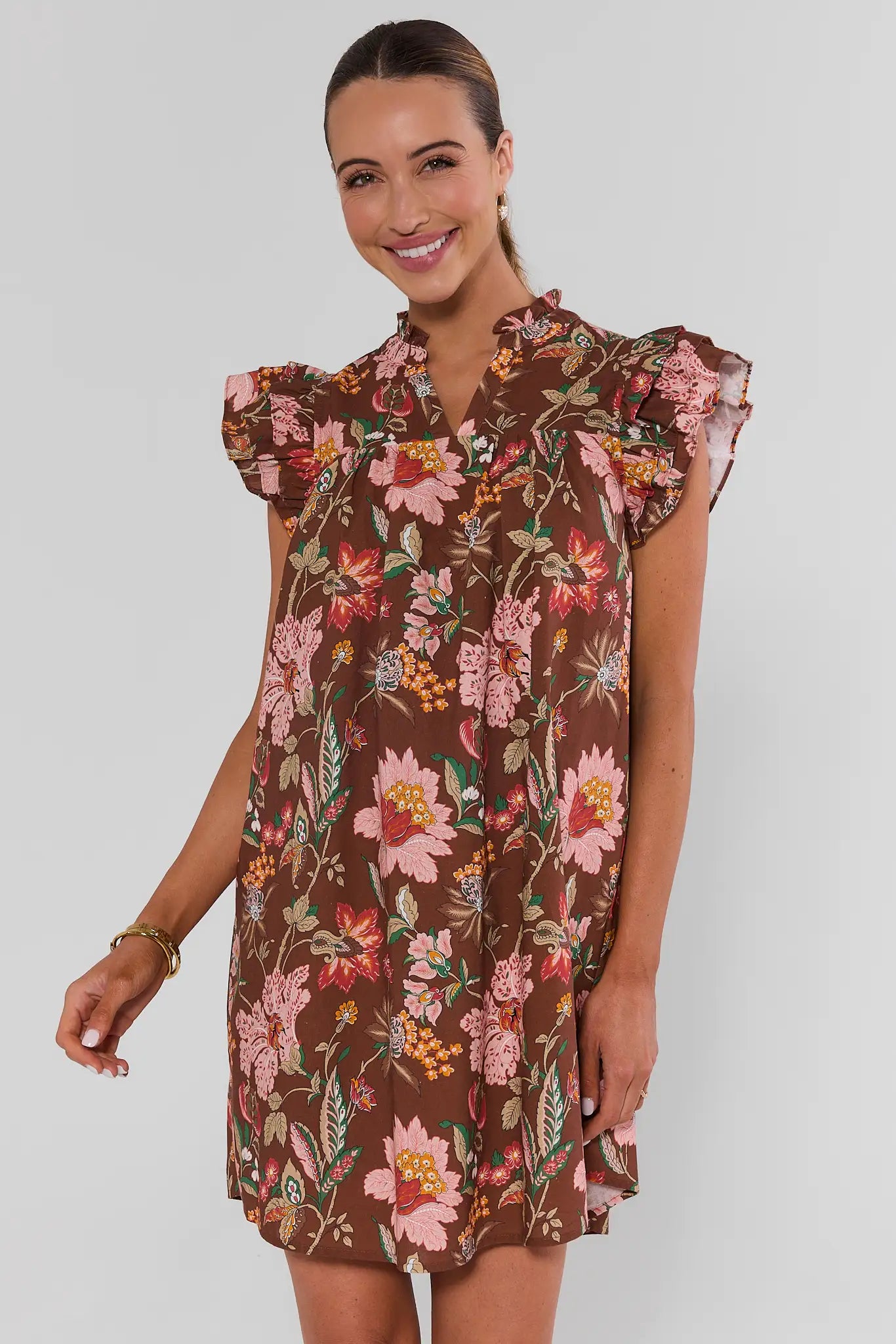 A woman modeling a brown short-sleeve floral dress with ruffled shoulders and a buttoned collar, photographed against a plain light gray studio background.
