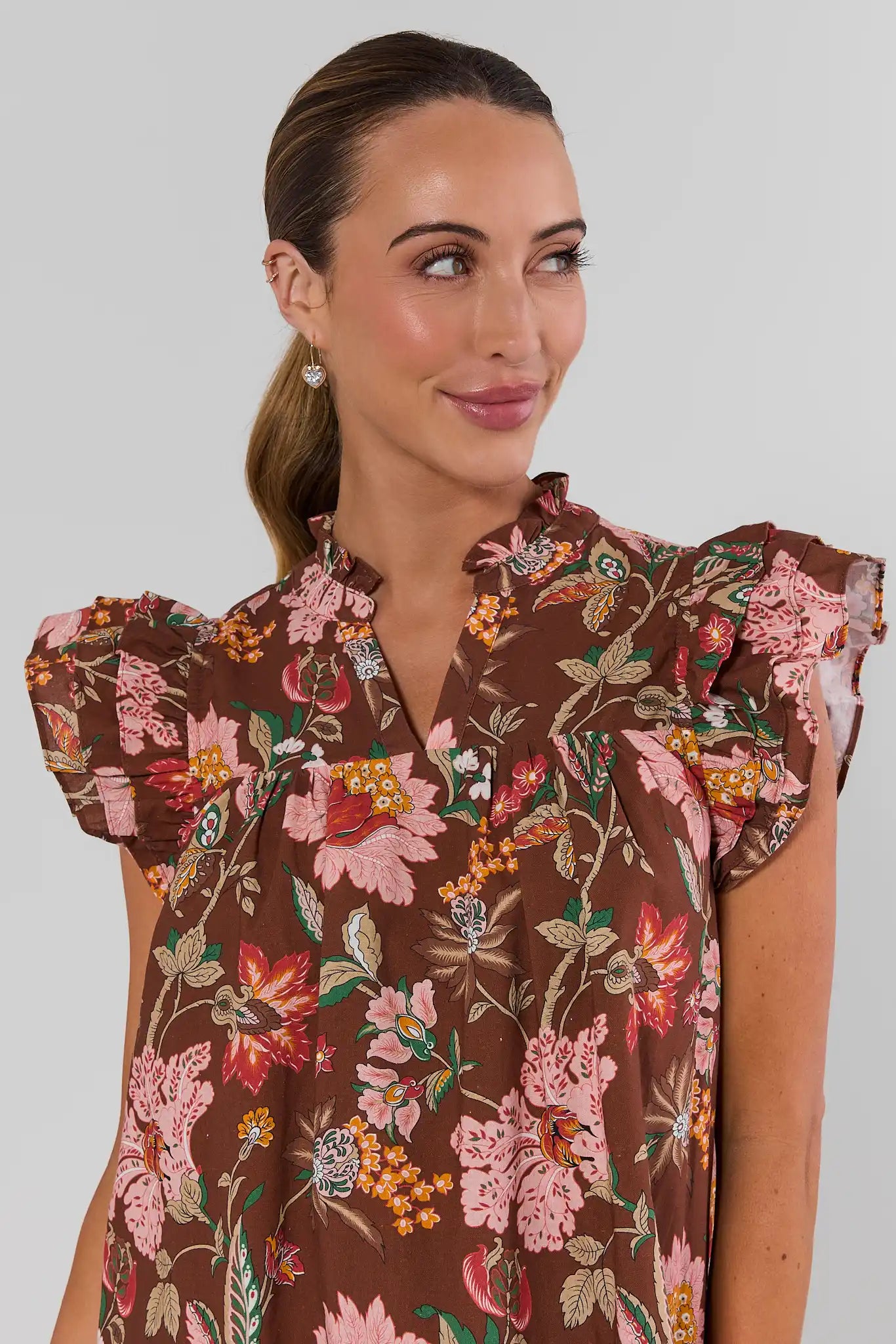A woman wearing a brown short-sleeve blouse with a vibrant pink, red, and green floral print, photographed against a solid light gray background. close up