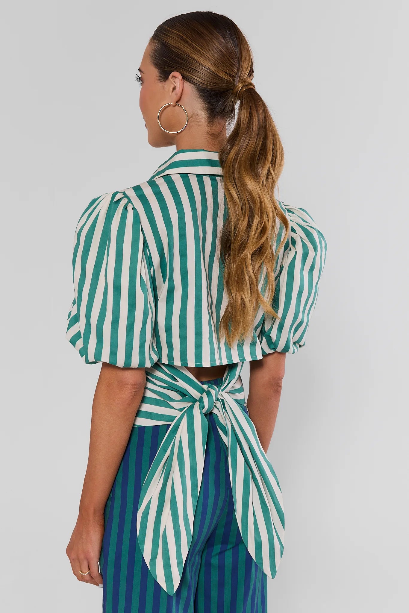 A woman wearing a green and white vertically striped crop top with puff sleeves and a tie-back waist, paired with blue and green striped pants