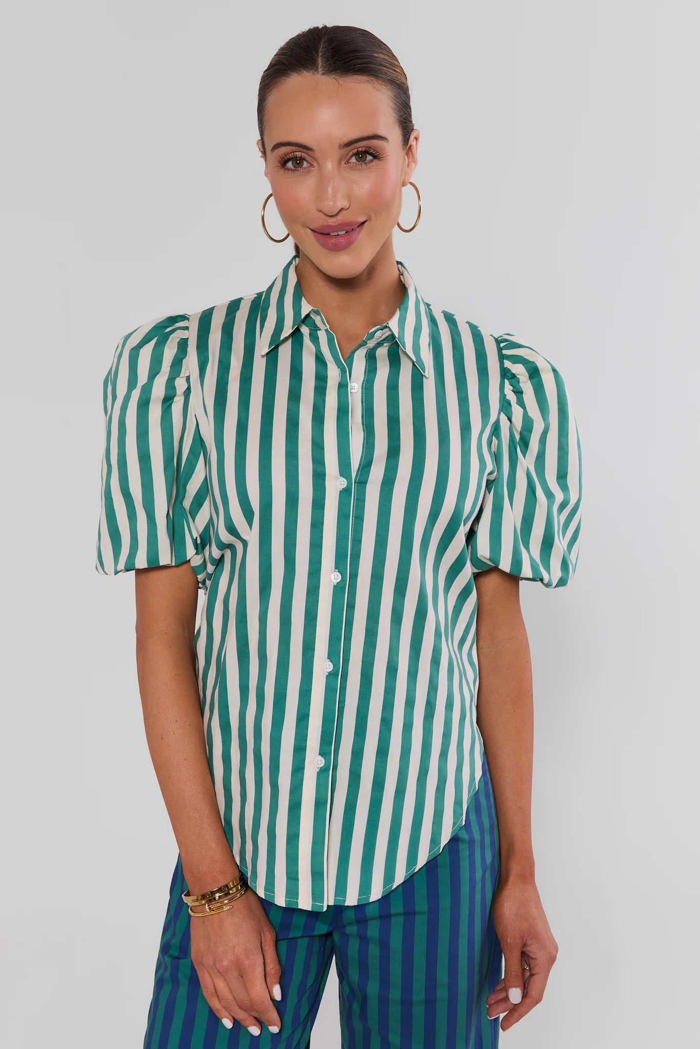 A woman modeling a short-sleeve button-up blouse with vertical green and white stripes and puffed shoulders, paired with blue-green striped trousers, against a light gray studio background.