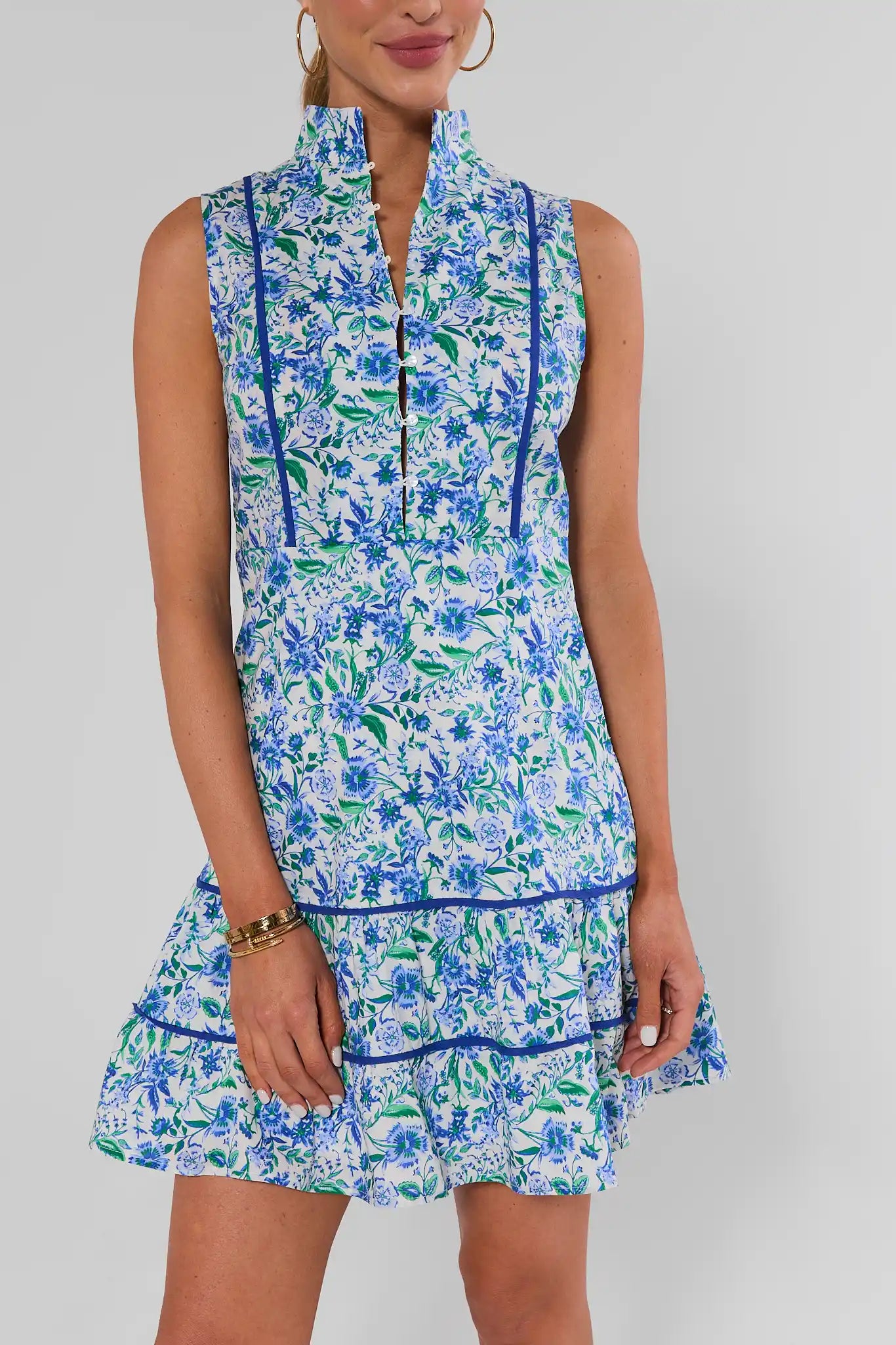 A sleeveless high-neck mini dress with a blue and green floral print on a white background, featuring royal blue piping details, shown on a model against a light gray studio background.