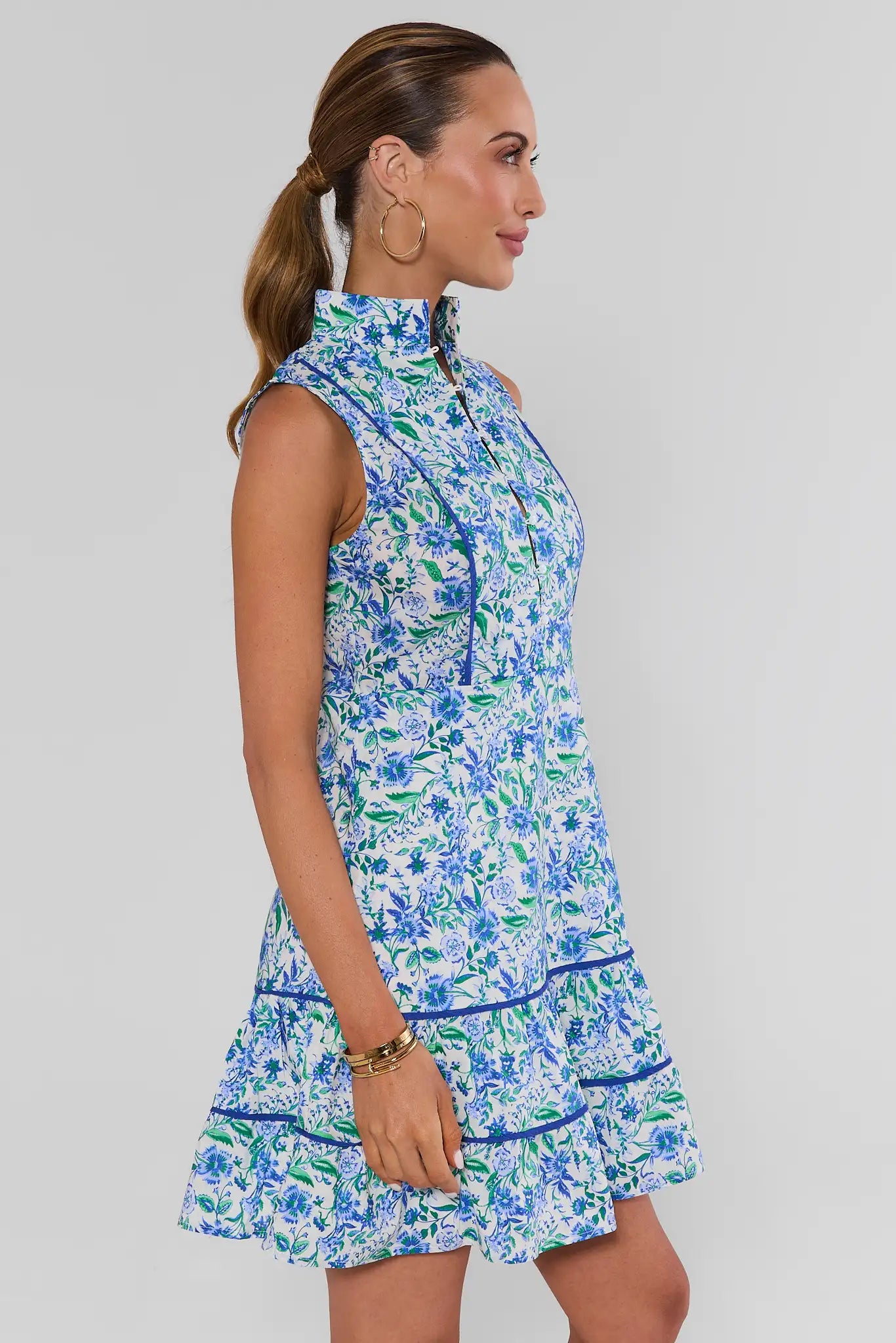 A woman shown in side profile wearing a sleeveless high-neck mini dress with a blue and green floral print on a white background, set against a plain light gray studio background.