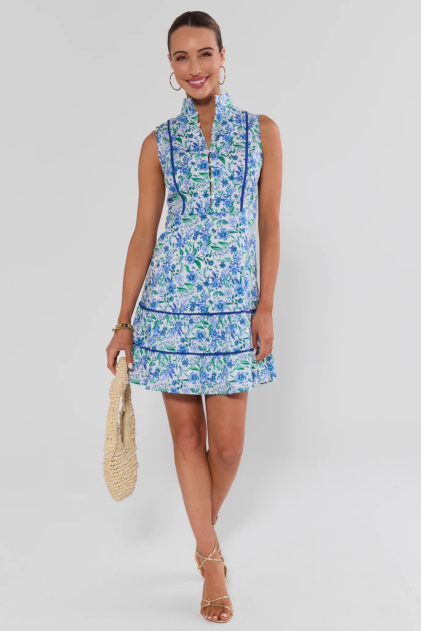 A woman modeling a sleeveless high-neck mini dress with a blue and green floral print, holding a woven bag against a gray studio background.