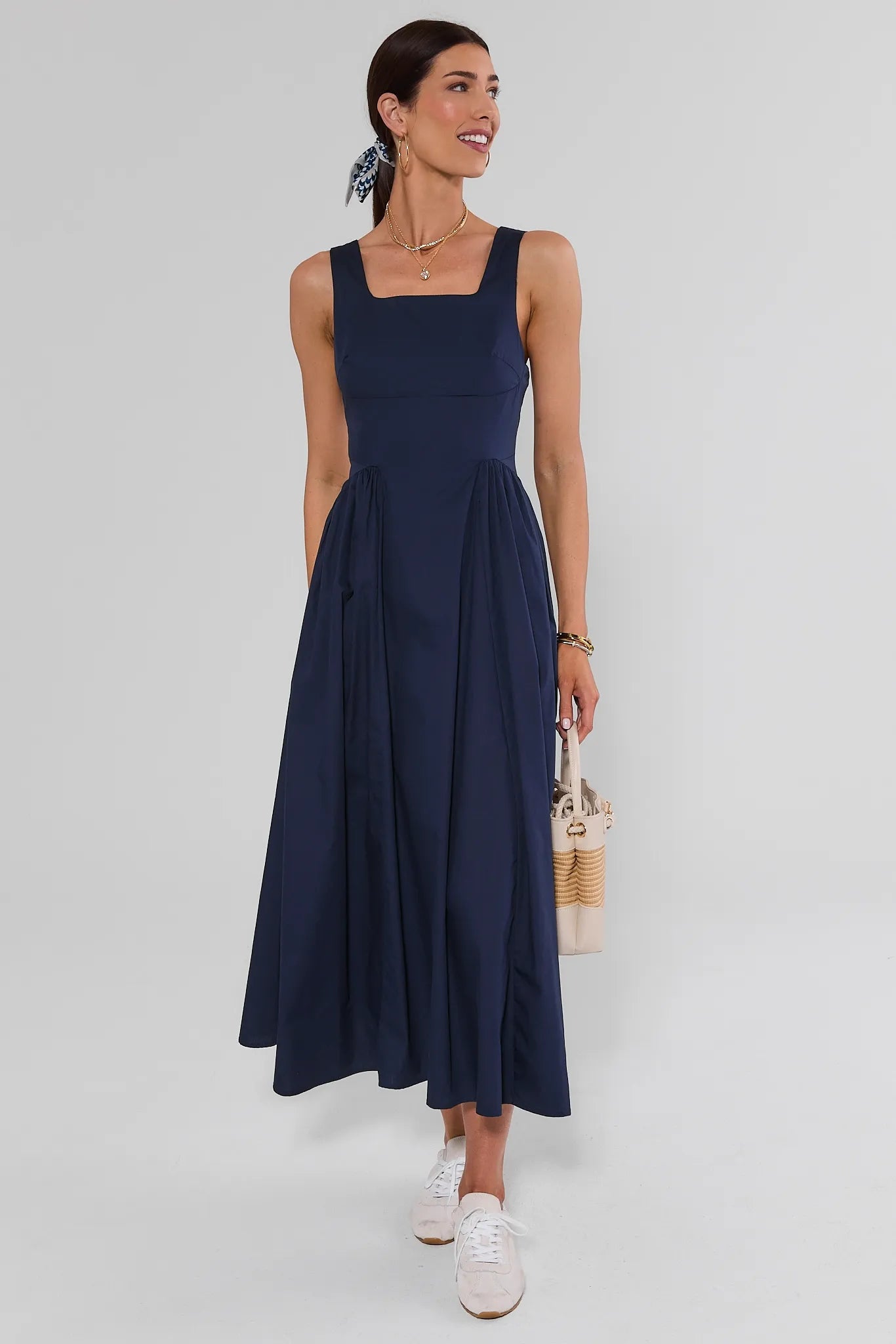 A woman modeling a sleeveless navy blue midi dress with a square neckline and a flared skirt, styled with white sneakers and a woven handbag against a light gray studio background.
