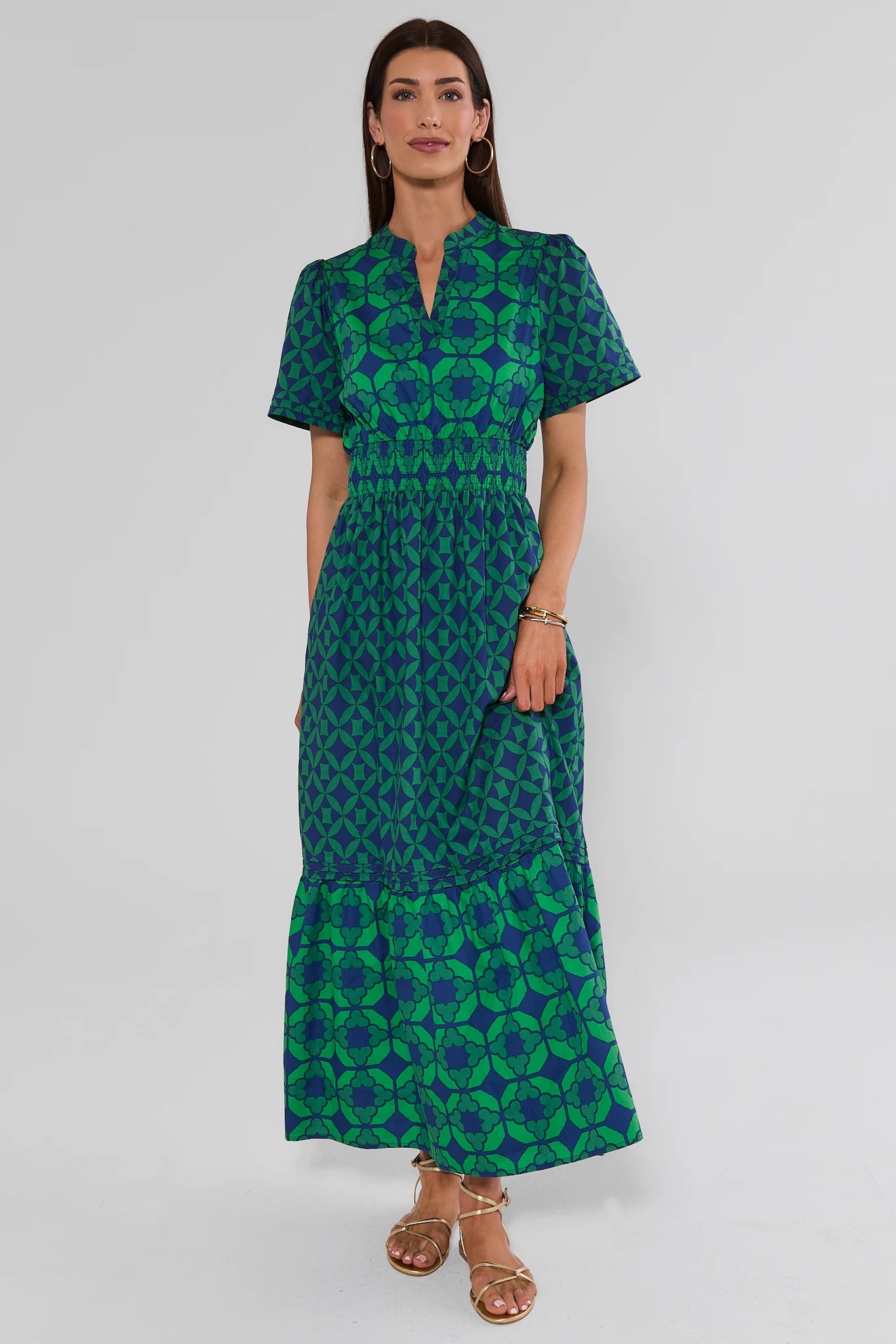 A woman standing wearing a green and blue patterned maxi dress with short sleeves and a smocked waist, on a white background