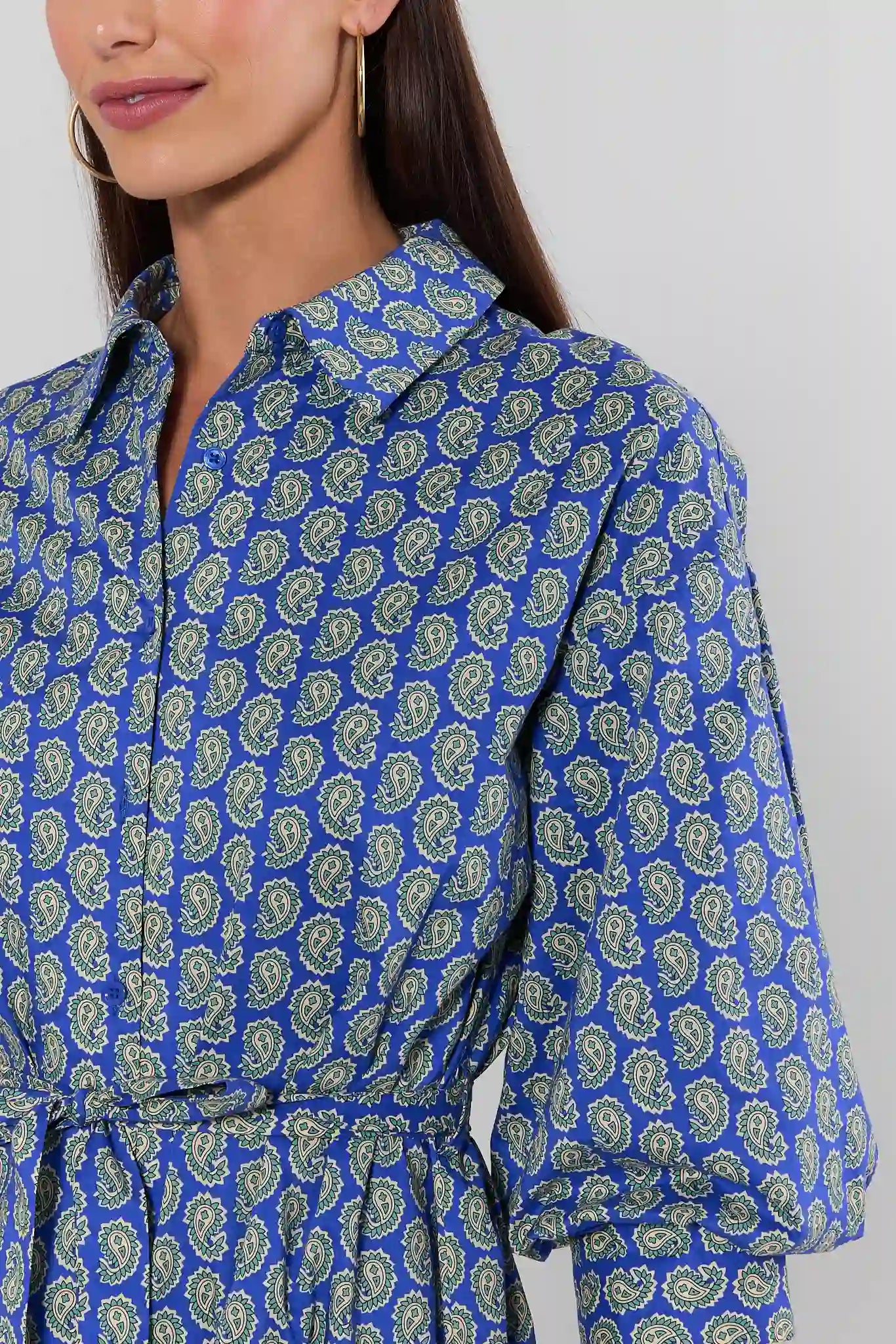 A close-up of a woman wearing a blue long-sleeve collared shirt dress with an all-over green paisley pattern and a matching waist tie.