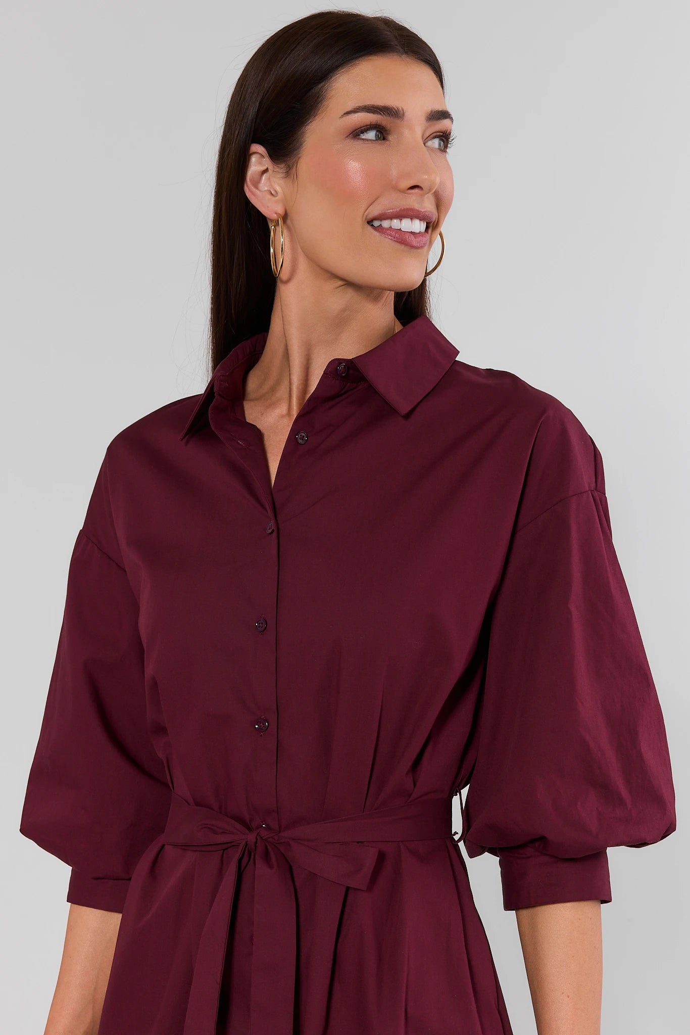 A woman wearing a dark burgundy button-up shirt dress with three-quarter puff sleeves and a matching tie waist belt, shown against a plain light background.