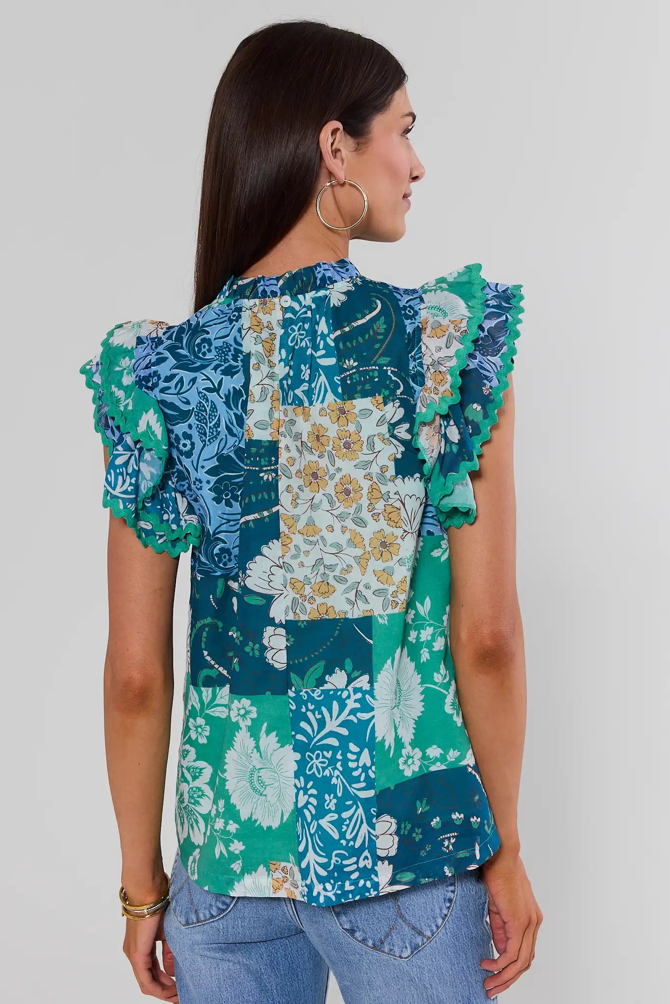 A woman turned with long hair wearing a colorful patchwork floral blouse with ruffled sleeves, on a white background