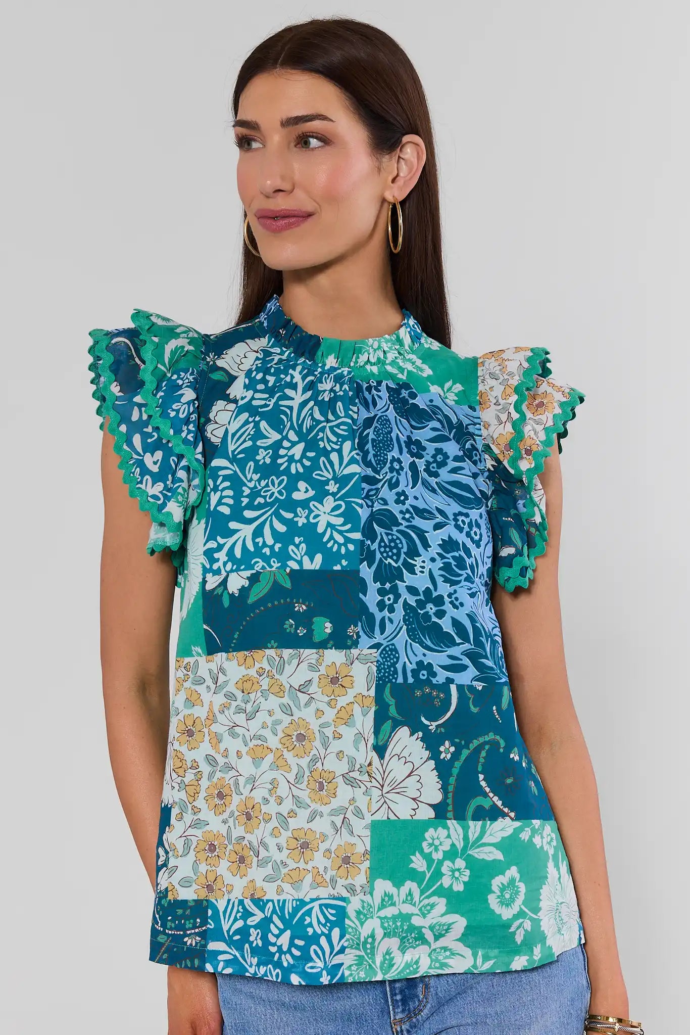 A woman with long brown hair wearing a colorful patchwork floral blouse with ruffled sleeves, with a white background