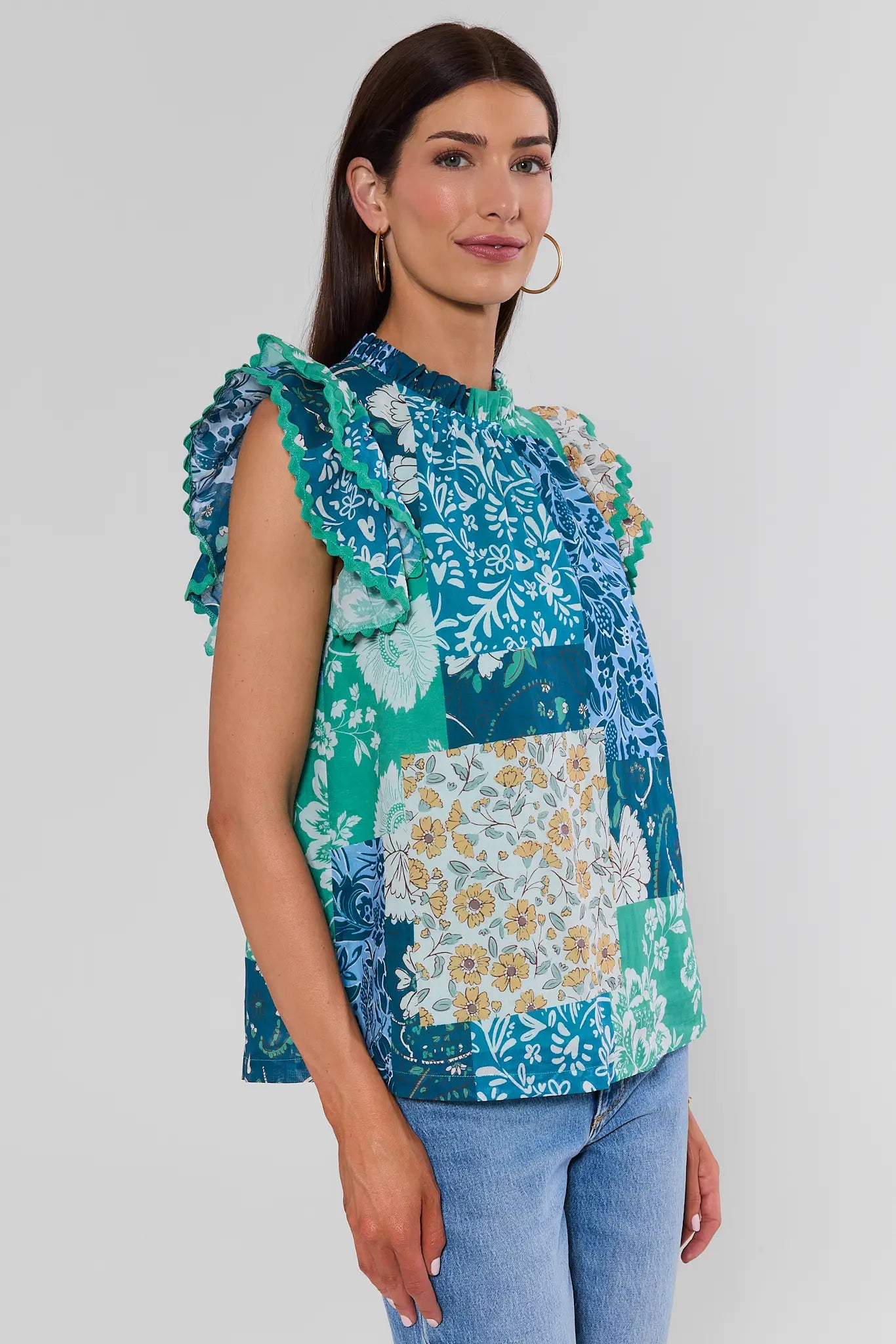 A woman modeling a sleeveless patchwork blouse with ruffled shoulders, featuring blue, green, and floral patterns, paired with light blue jeans against a light gray studio background.
