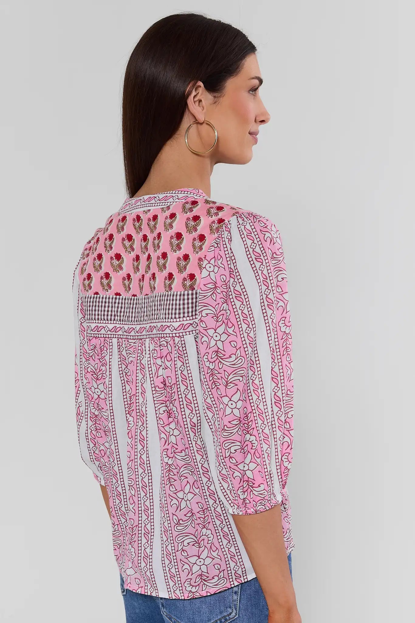 A woman shown from the back wearing a pink and white patterned blouse with floral embroidery and a gingham band across the upper back, paired with blue jeans against a light gray studio background.