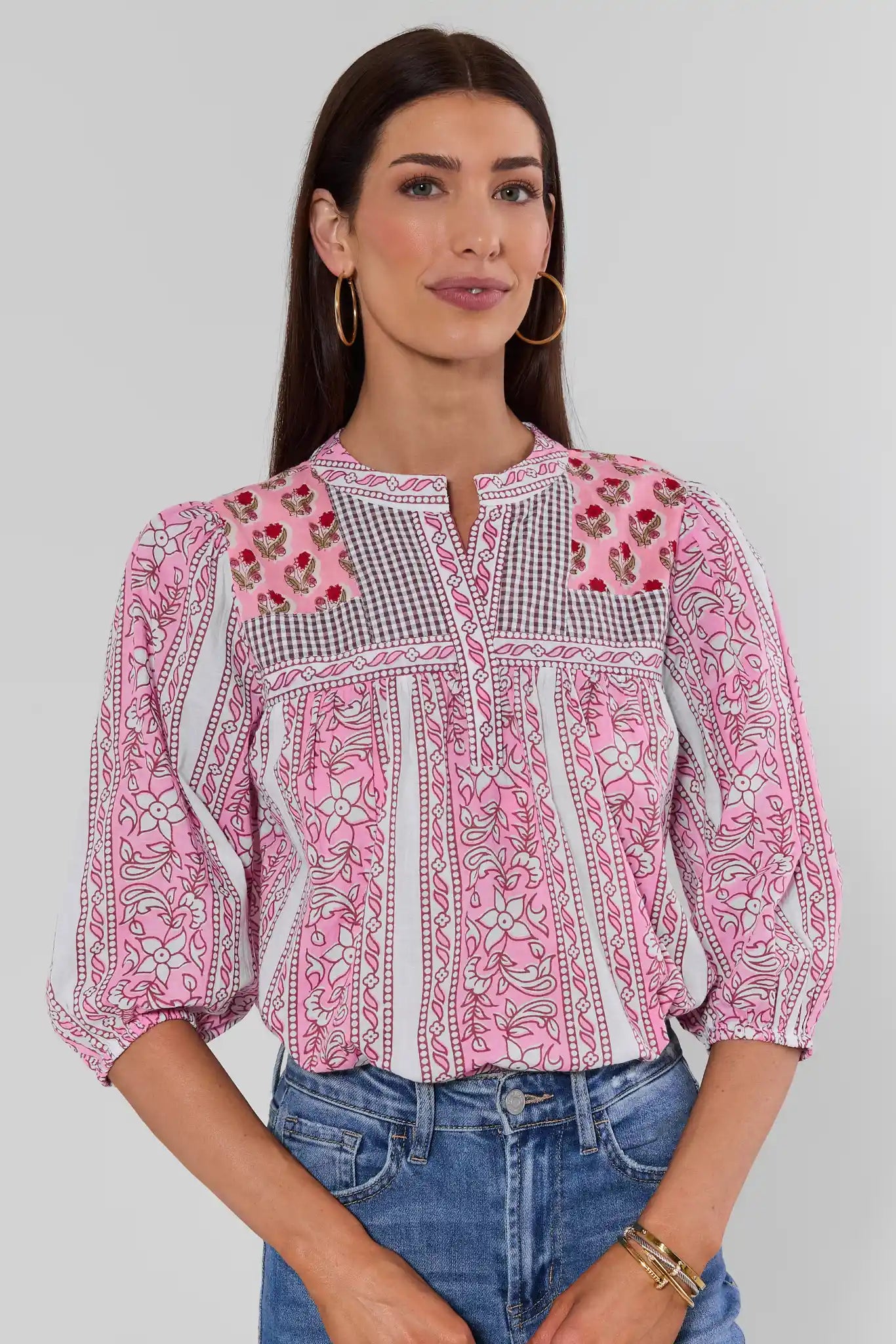 A woman wearing a pink embroidered top with white and black gingham panels, floral embroidery, and geometric borders, photographed against a studio background.