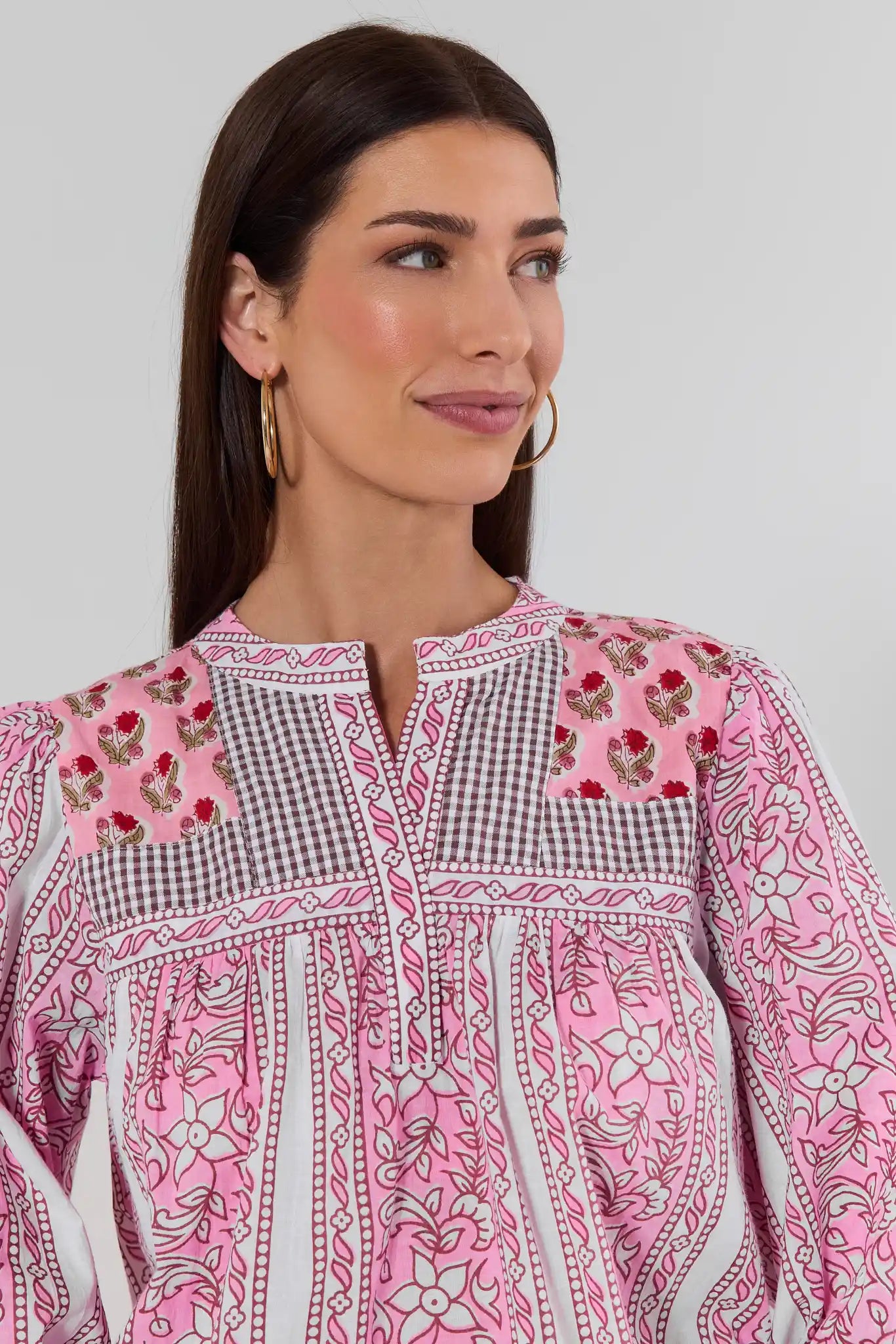 A woman wearing a pink embroidered top with white and black gingham panels, floral embroidery, and geometric borders, photographed against a light gray studio background.