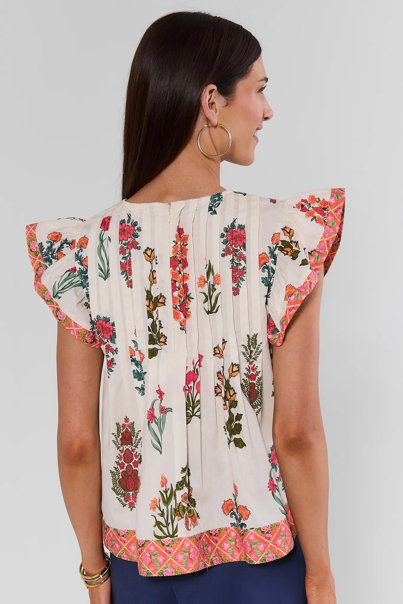 A woman shown from the back wearing a cream-colored blouse with colorful floral prints and ruffled flutter sleeves, paired with navy blue pants, against a light studio background.