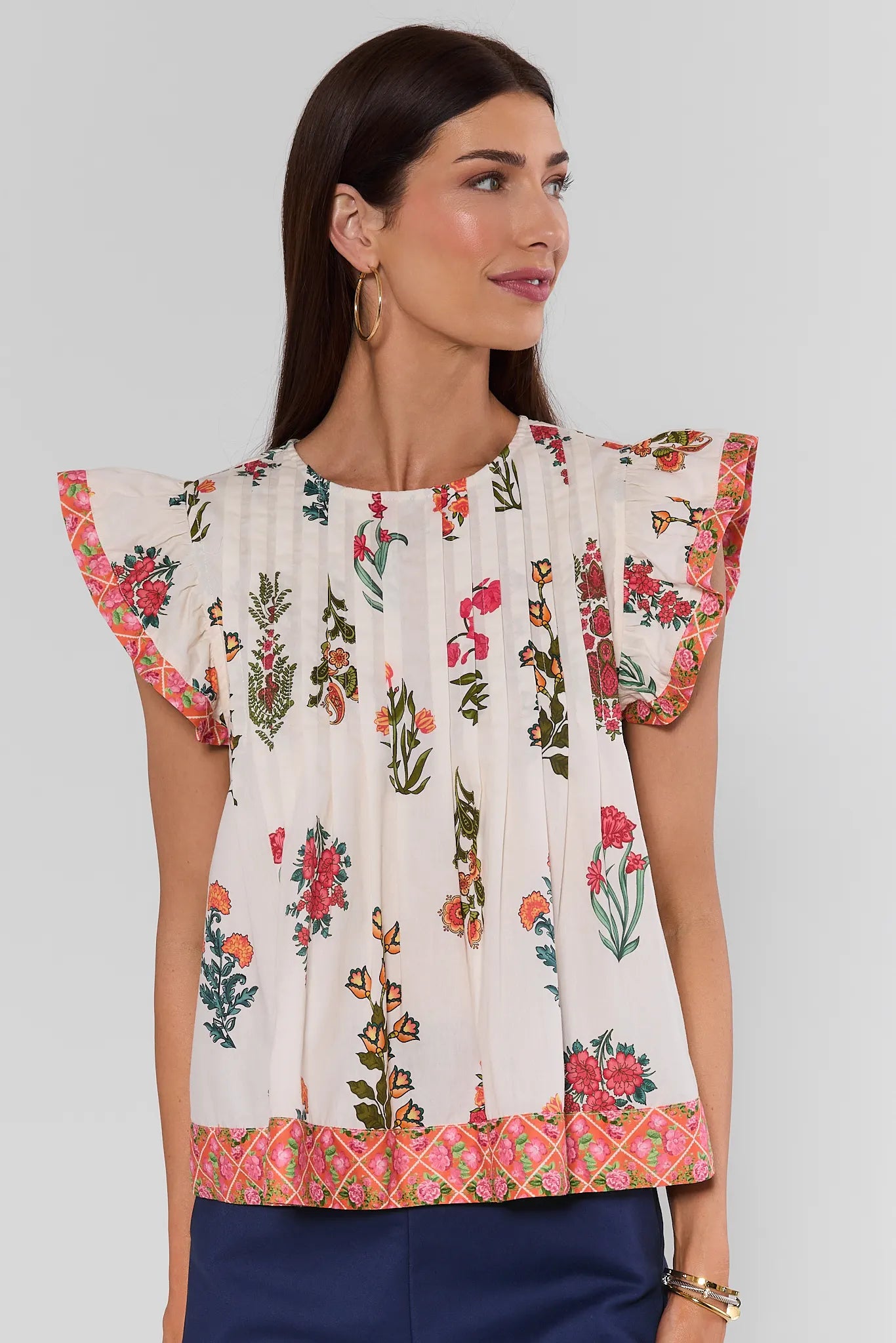 A woman wearing a white short-sleeve blouse with colorful floral prints and patterned ruffled sleeves, paired with navy blue pants, against a light gray studio background.