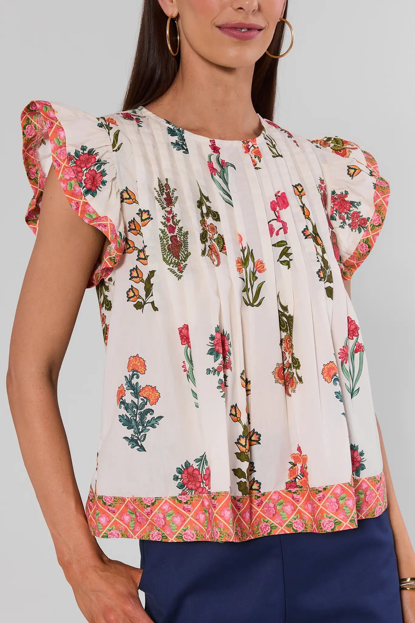 A woman wearing a short-sleeve blouse with a cream base and a colorful floral and butterfly print, paired with navy blue pants. The blouse features ruffled sleeves and a patterned hem.