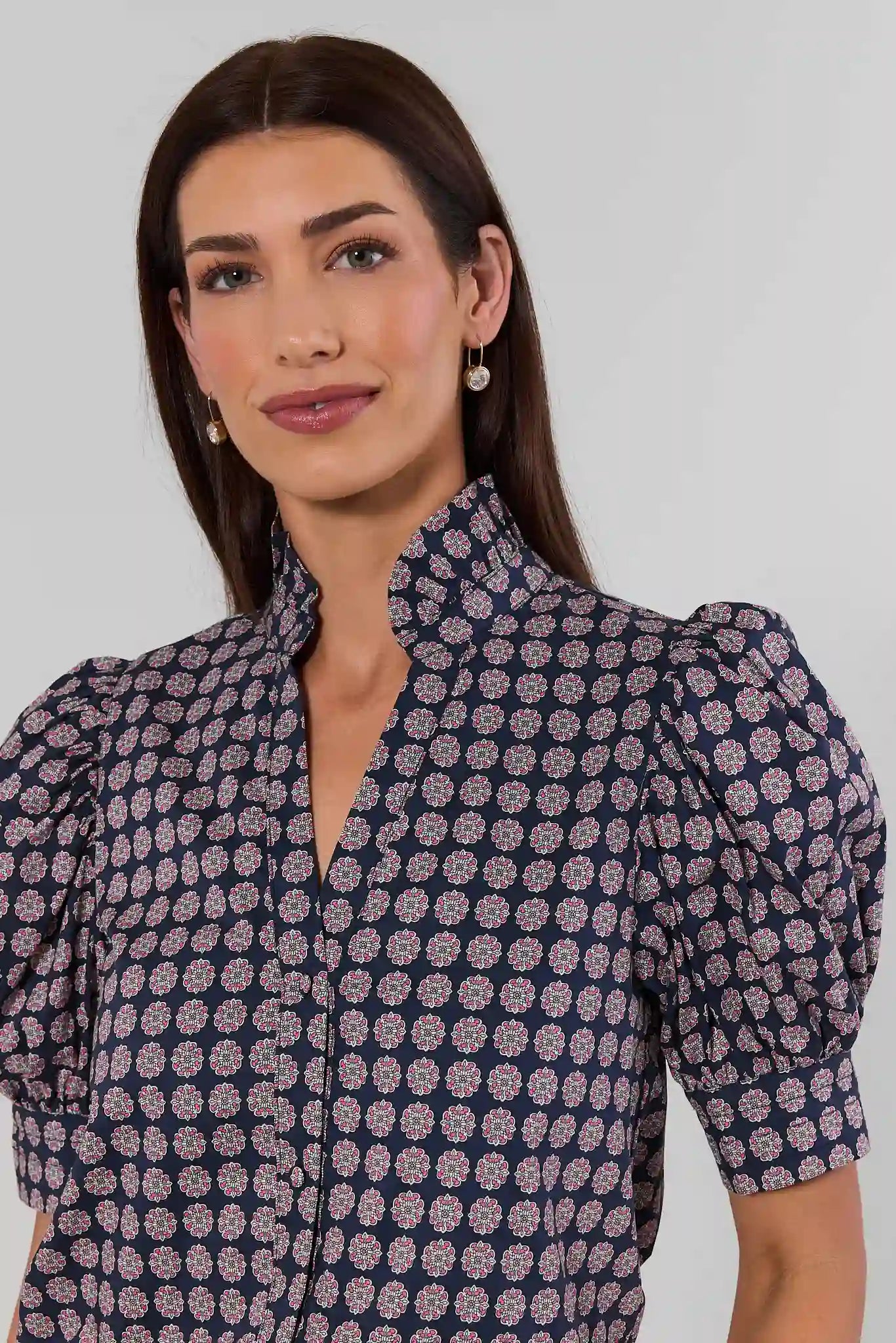 A woman wearing a navy blue blouse with a dense pink and white floral pattern, featuring a high mock neck and puffed short sleeves, set against a plain light gray background.