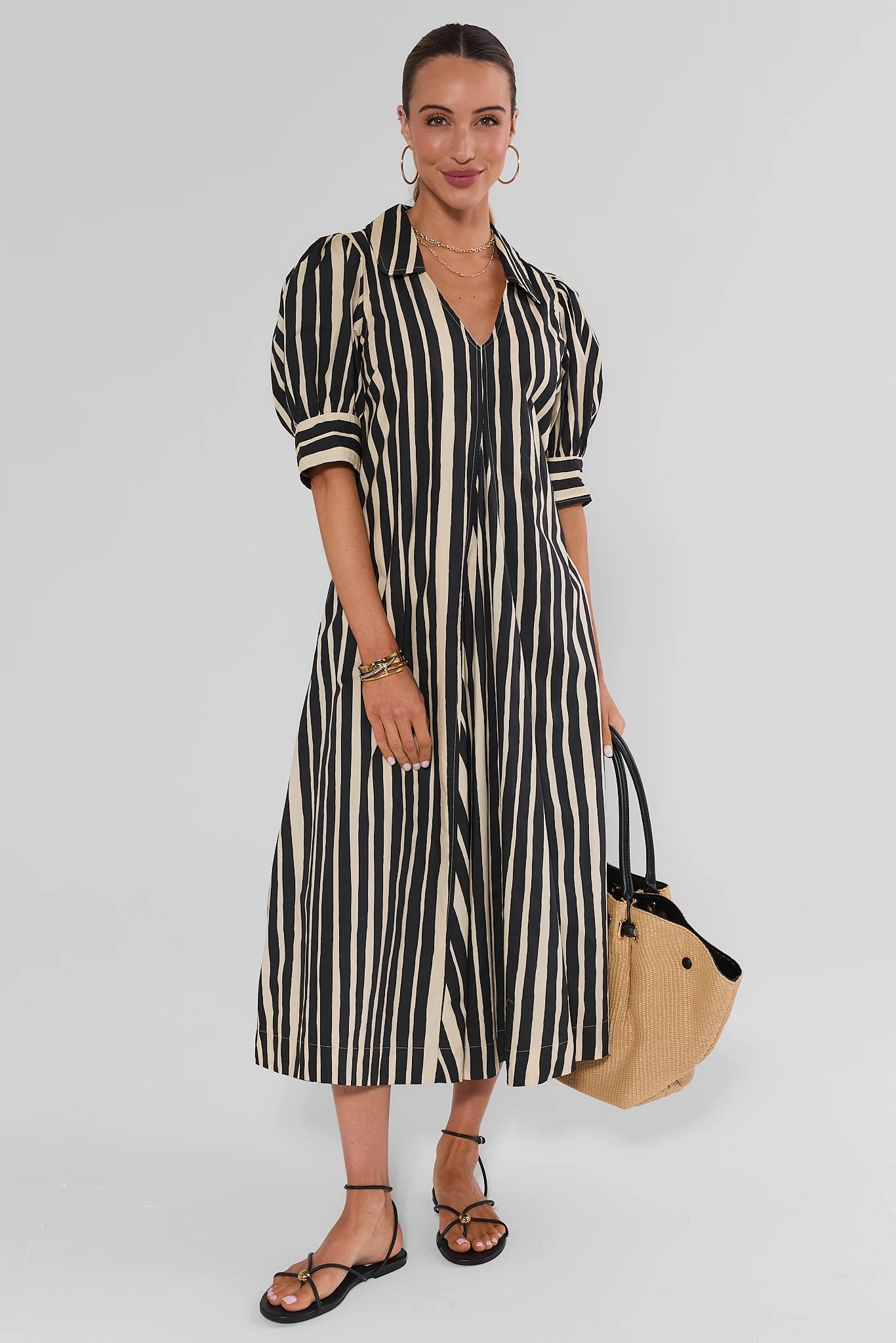A woman models a black and cream vertical striped midi shirt dress with puff sleeves, holding a beige tote bag against a light gray studio background.