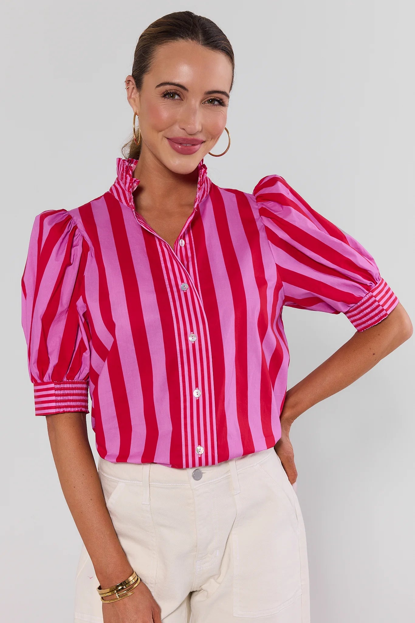 A woman wearing a vibrant pink and red vertically striped blouse with puff sleeves and a high ruffled collar, on a white background.