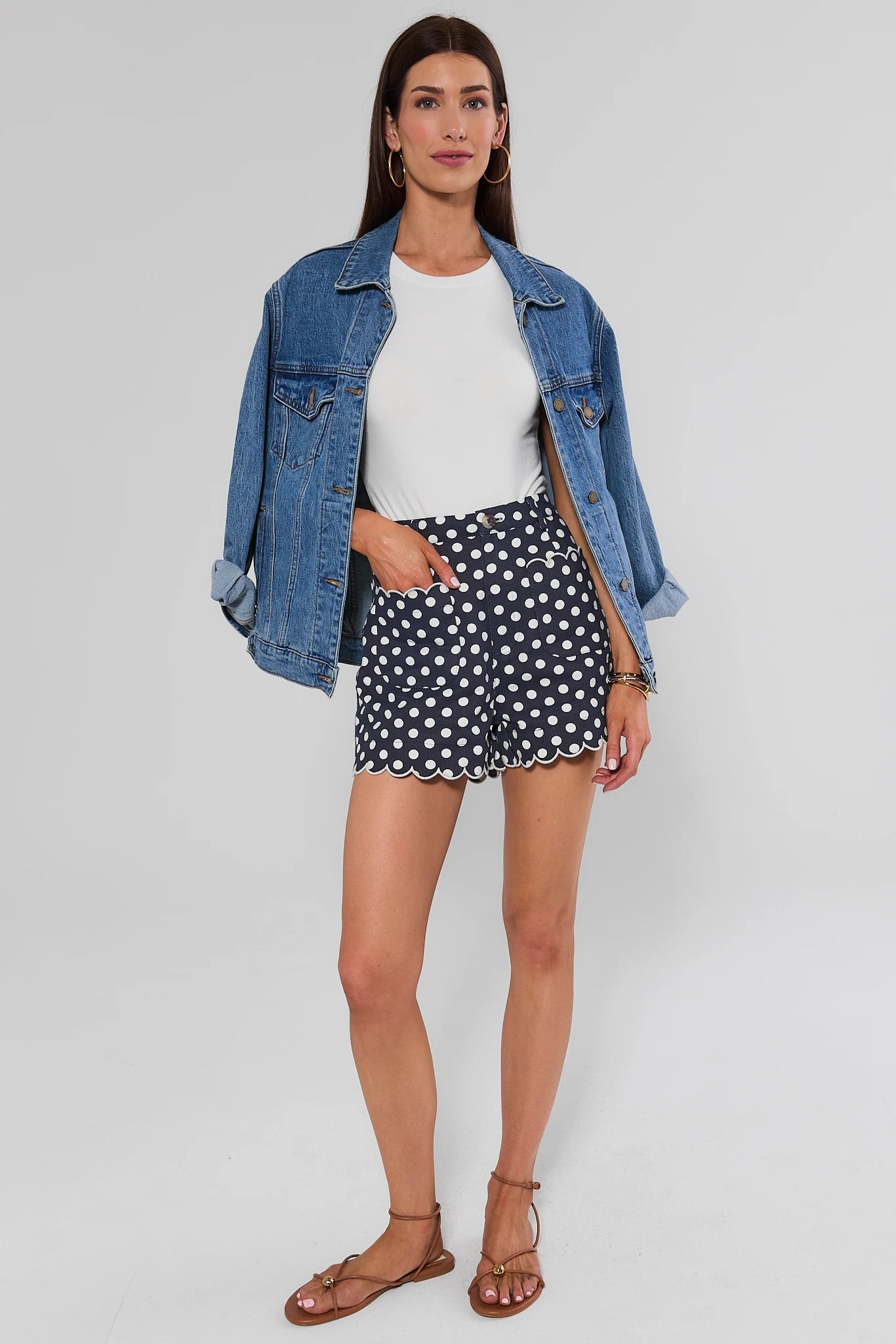 A woman modeling a white top and denim jacket and navy blue shorts with white polka dots and scalloped hems, against a light background.