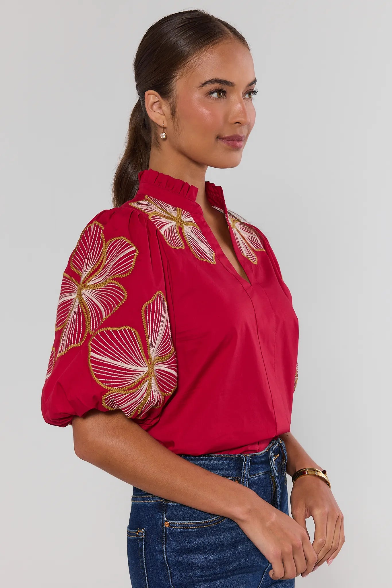 woman wearing a bright red blouse with large white and gold floral embroidery on the bodice and sleeves, paired with a high ruffled collar and puff sleeves turned to side