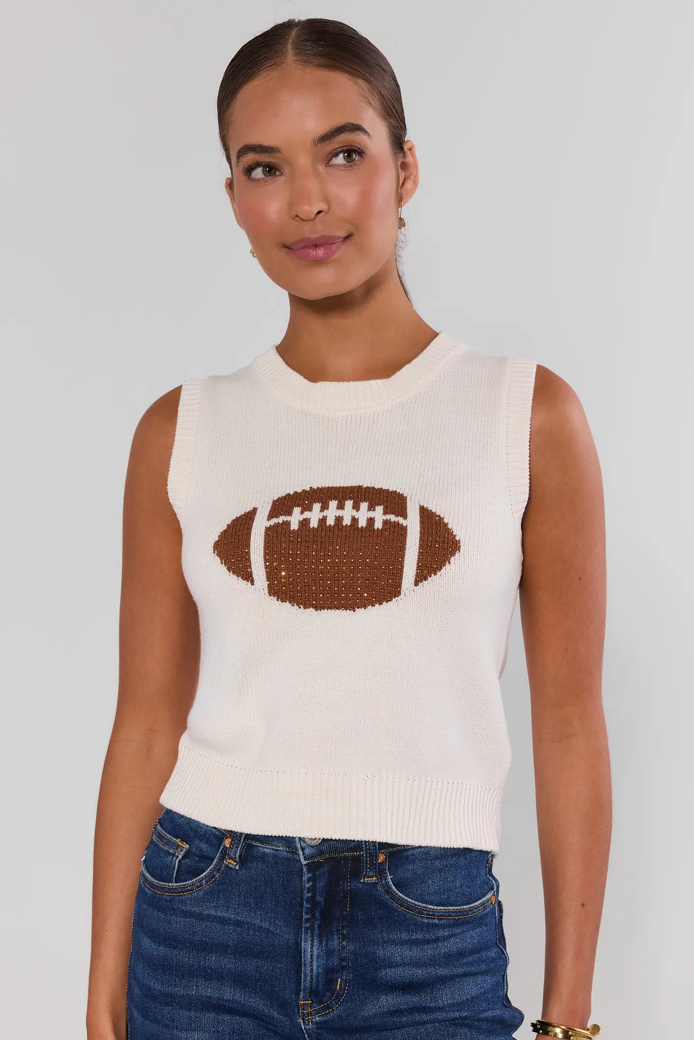 A woman wearing a sleeveless cream knit sweater vest with a brown football graphic on the front, paired with blue jeans, against a light gray studio background.