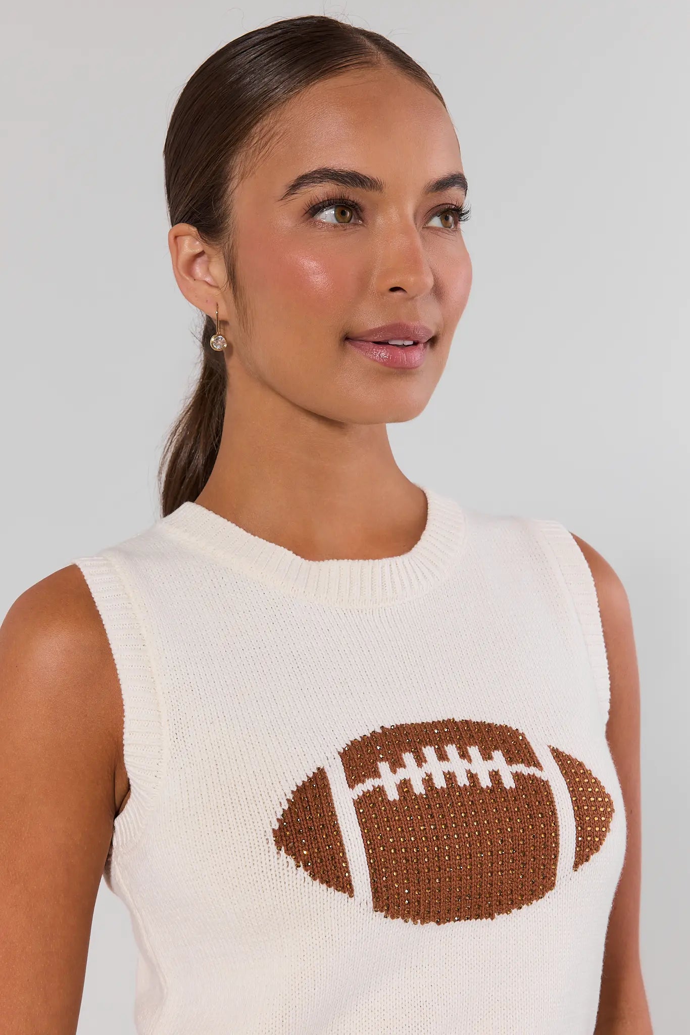 A woman wearing a cream knit sleeveless sweater with a brown and white football graphic on the front, set against a plain light gray background close up