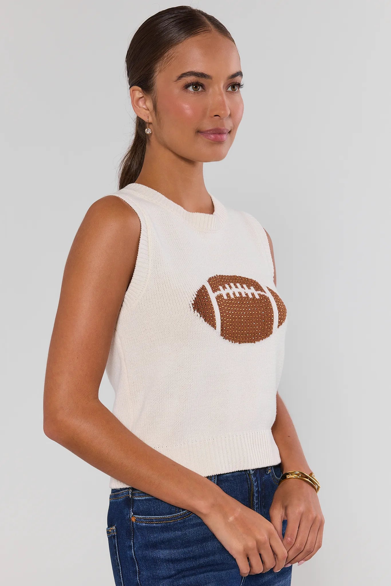 A woman modeling a sleeveless white top with a brown football graphic on the chest, paired with dark blue jeans and beige sneakers, against a light gray studio background turned to side