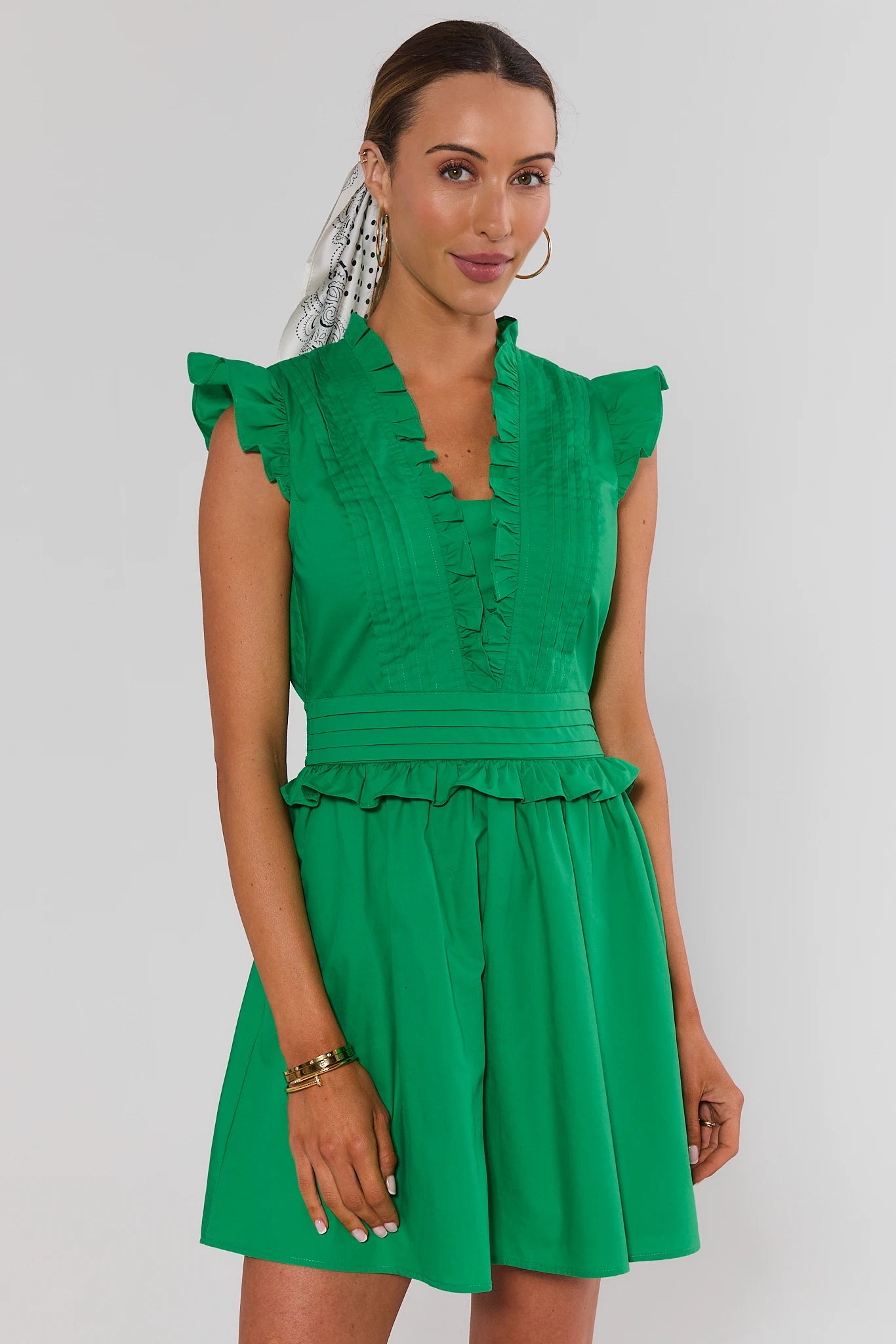 A woman modeling a bright green mini dress with ruffled cap sleeves, a ruffled V-neckline, a defined waist, and a flared skirt, photographed against a light gray studio background.
