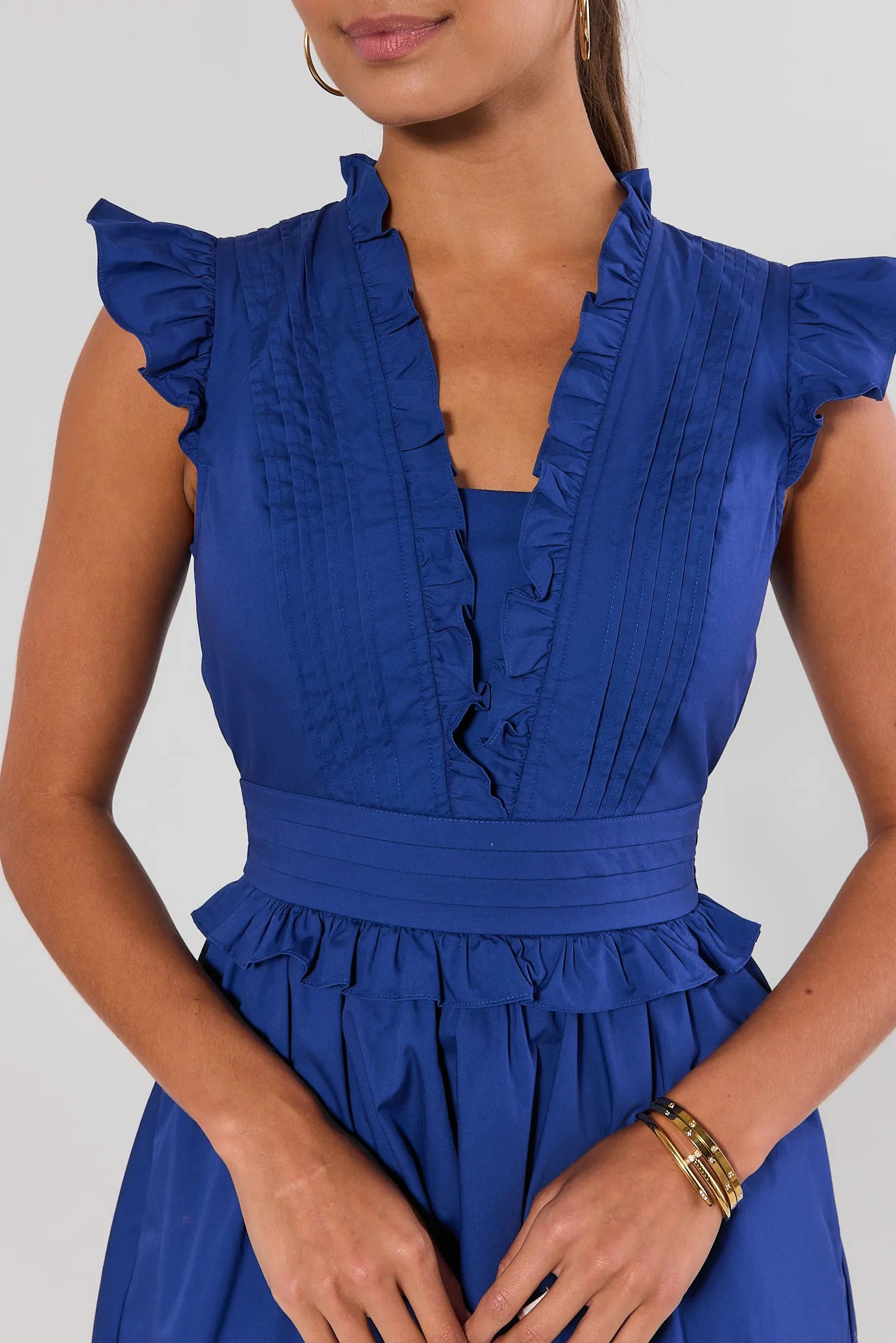 Close up of a woman modeling a royal blue sleeveless mini dress with ruffled trim, cap sleeves, a gathered waist, and a flared skirt against a light gray studio background.
