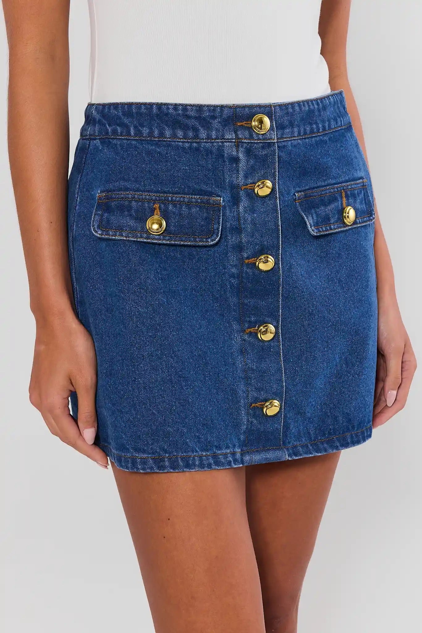 Close up of a model wearing a blue denim mini skirt with gold-tone button details and flap pockets, paired with leopard-print flat shoes, against a light gray studio background.