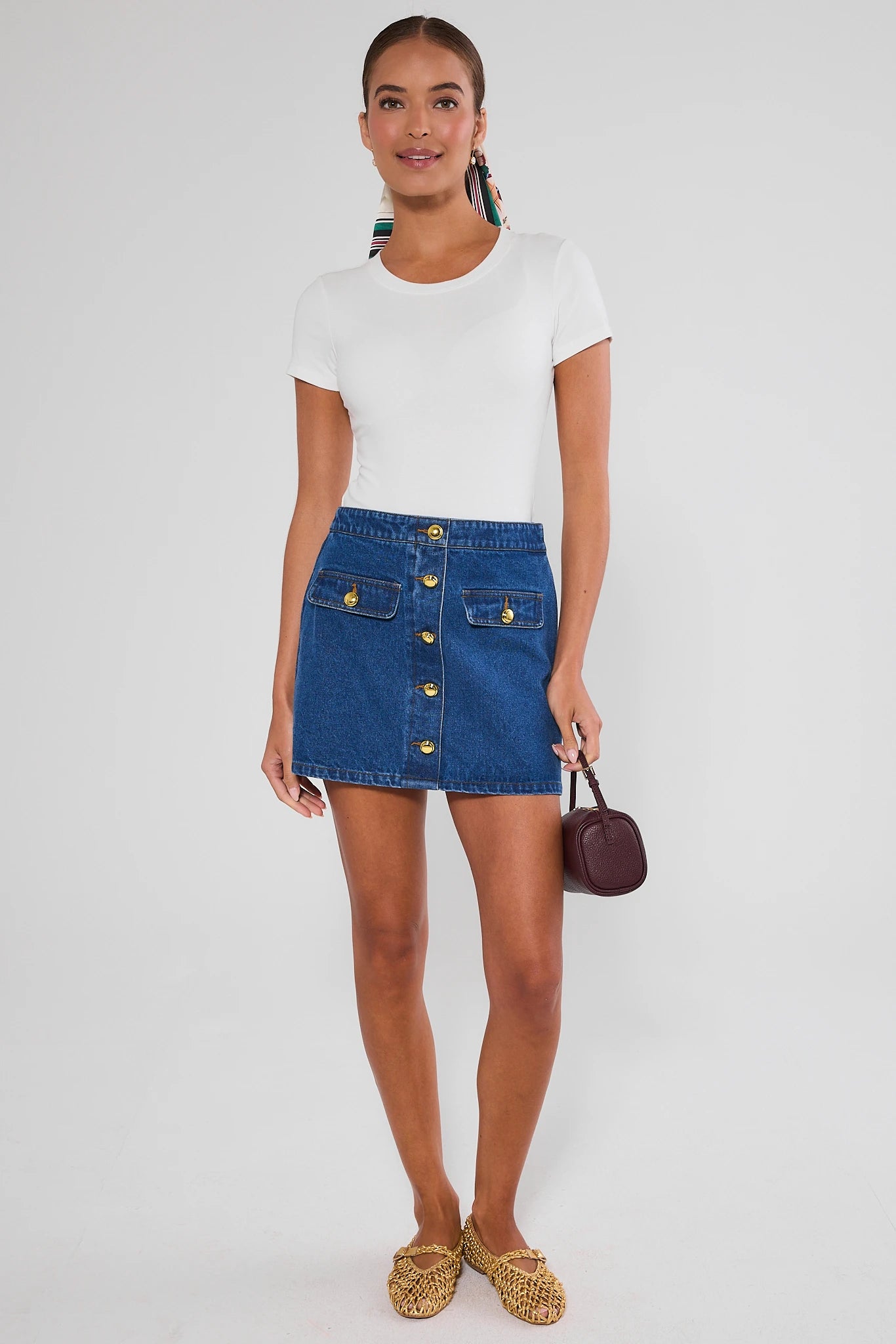 A model wearing a white top and blue denim mini skirt with gold-tone button details and flap pockets, paired with gold flats, against a light gray studio background.