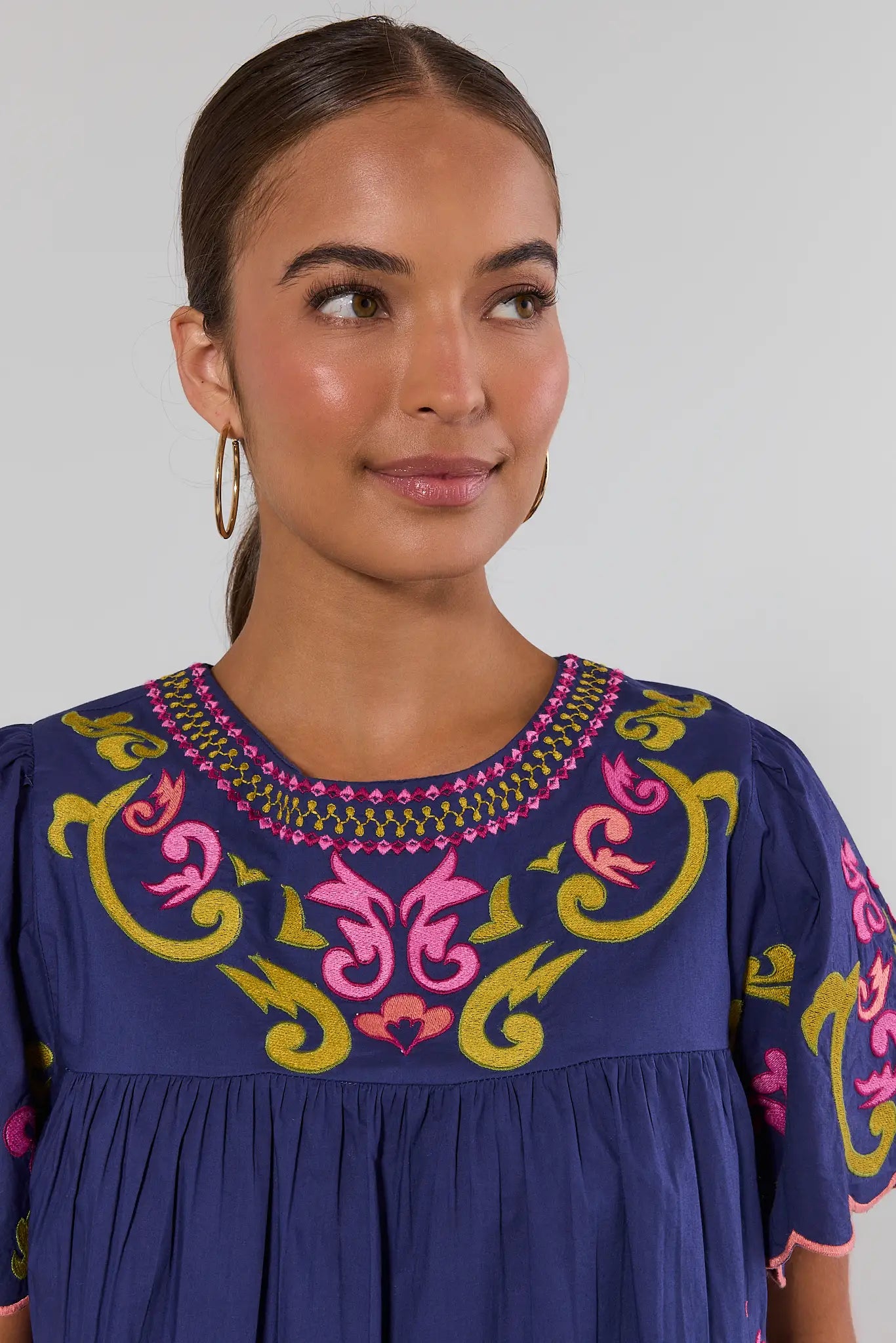 A woman wearing a navy blue blouse with colorful pink, yellow, and green embroidery and scalloped short sleeves, paired with light blue jeans, against a light gray background close up