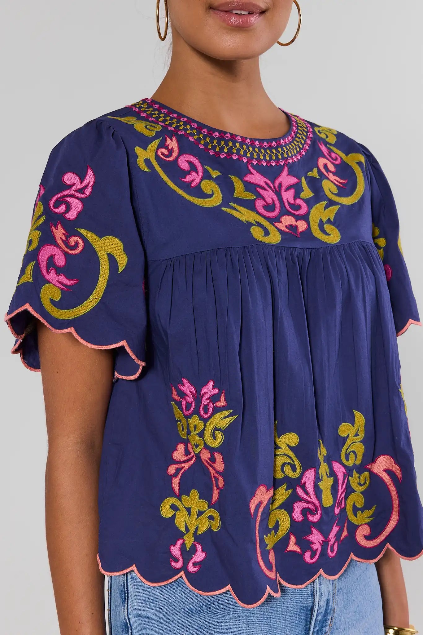 close up A woman wearing a navy blue blouse with colorful pink, yellow, and green embroidery and scalloped short sleeves, paired with light blue jeans, against a light gray studio background.